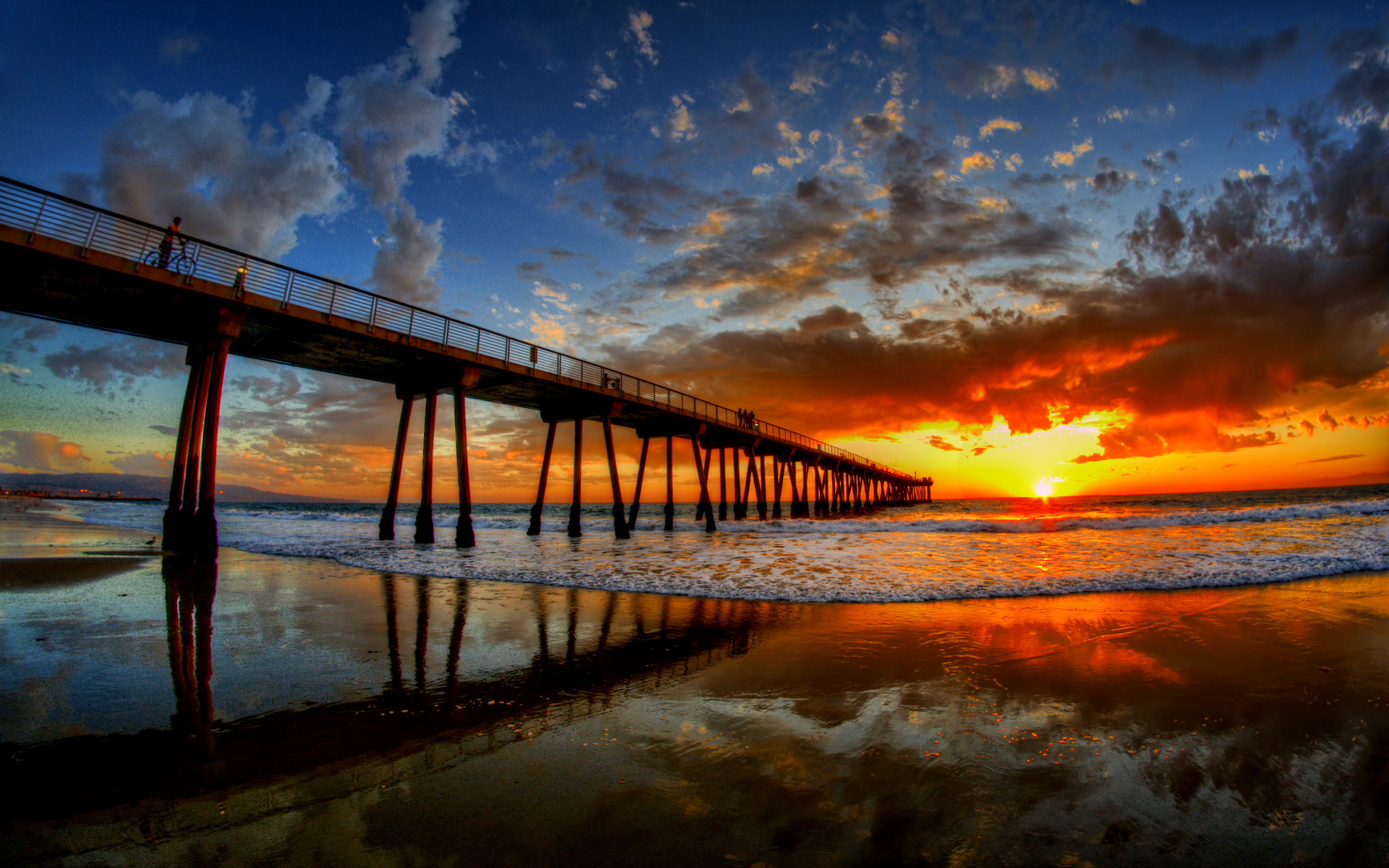 A breathtaking sunset over the ocean with a pier in the foreground. The sky is ablaze with vibrant orange and red hues, reflected in the wet sand beneath the pier