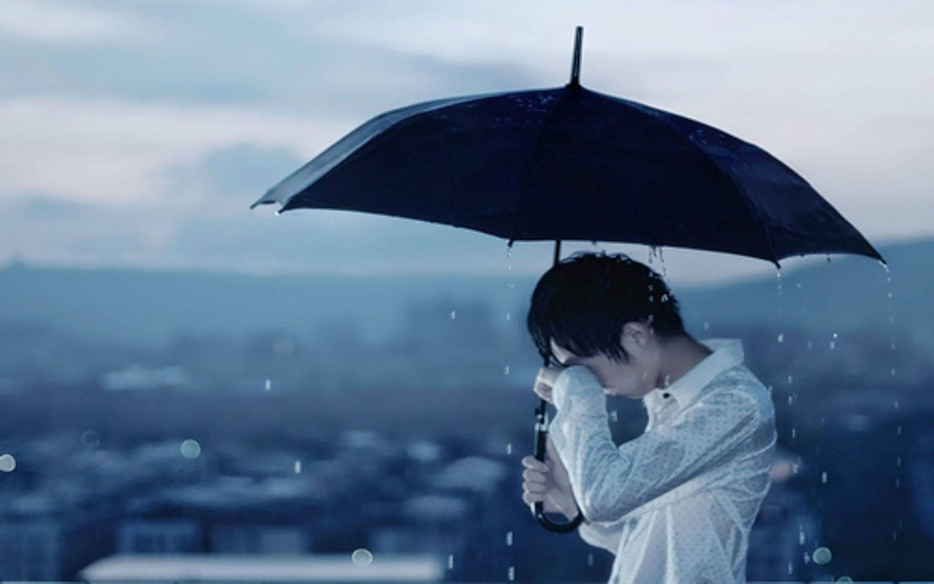 A person stands with their head down, holding a black umbrella in the rain. The city skyline is blurred in the background