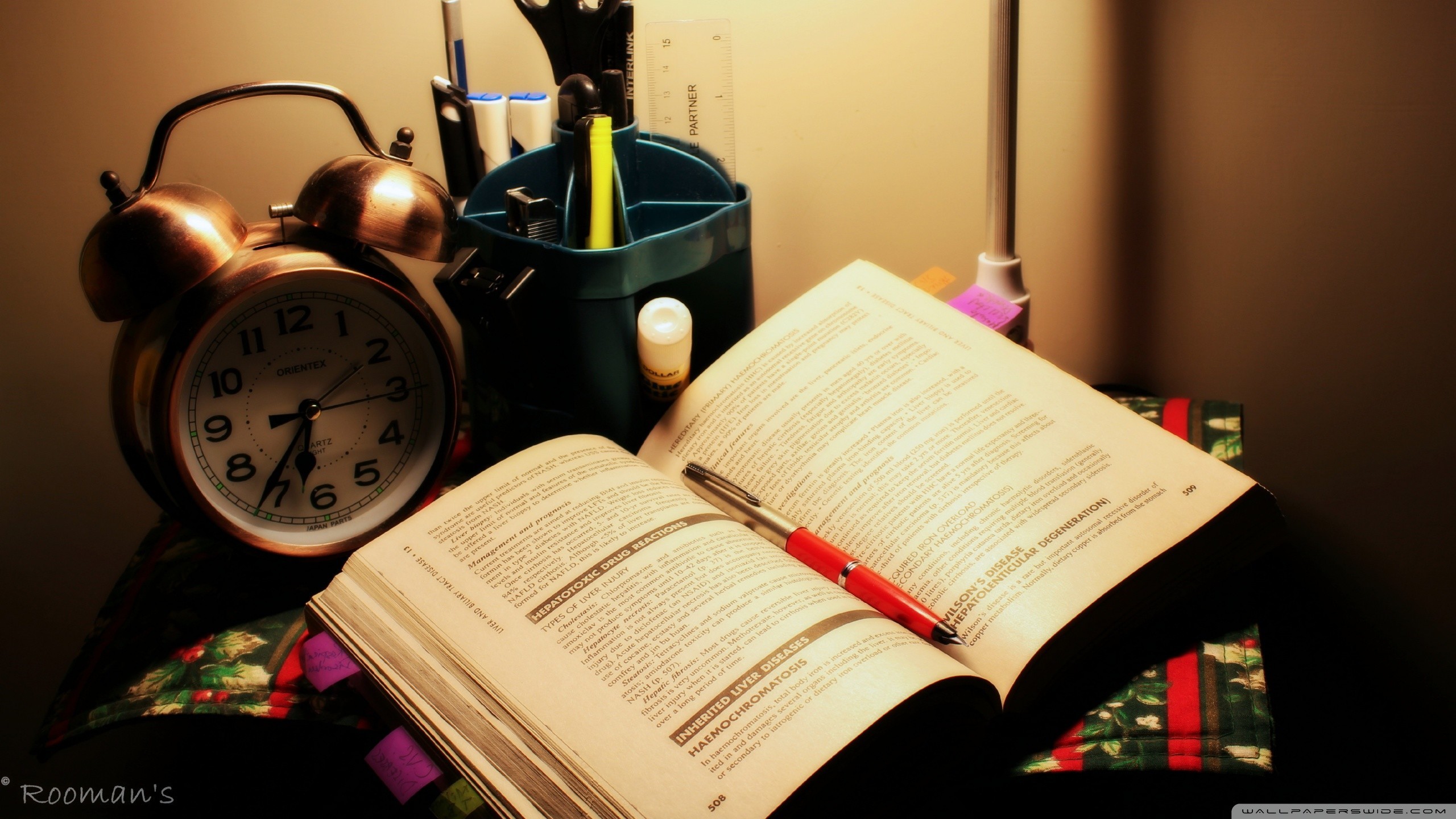 Close-up of an open textbook, a red pen, an alarm clock, and a desk organizer, illuminated by a warm lamp, creating a cozy study atmosphere