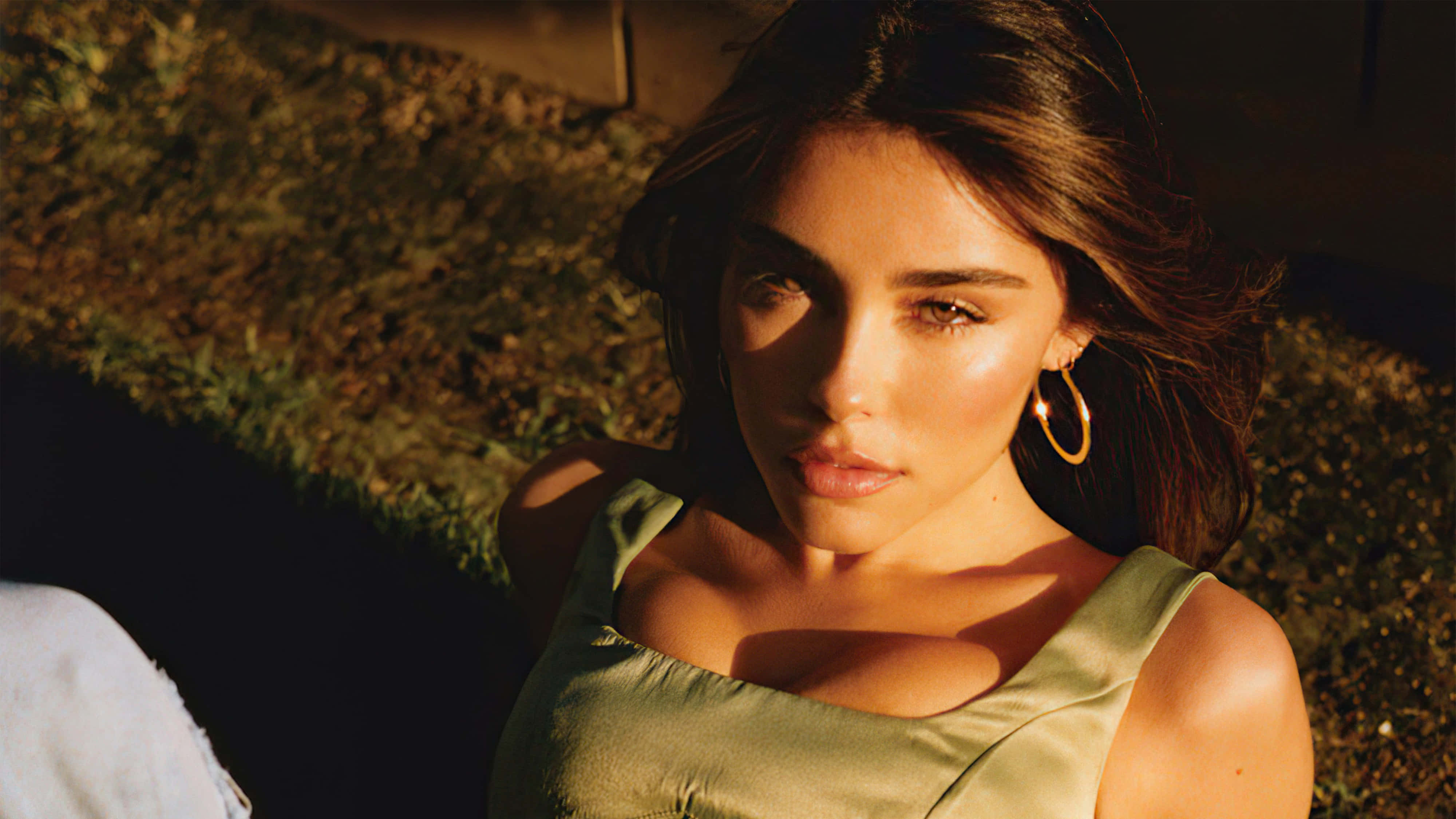 A young woman with long brown hair and gold hoop earrings, wearing a light green top, is seen from the chest up, looking directly at the camera. She is outdoors, and the sunlight casts shadows on her face and body