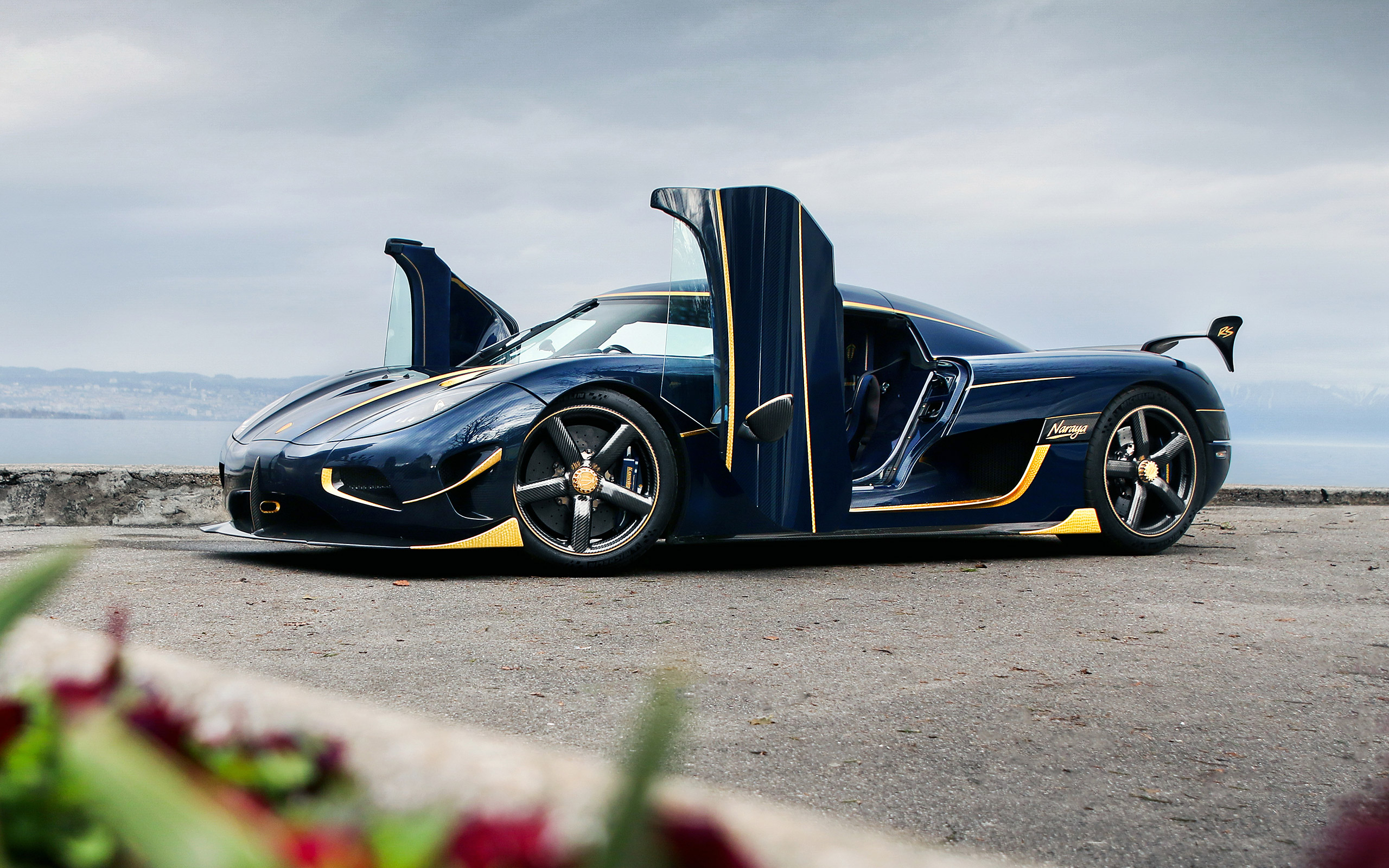 A dark blue Koenigsegg Agera RS with gold accents, parked by a lake with its dihedral synchro-helix actuation doors open