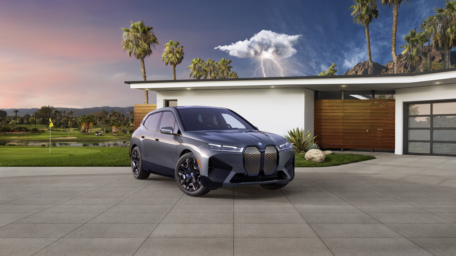 A gray BMW iX electric SUV parked in a driveway in front of a modern house, with a golf course in the background