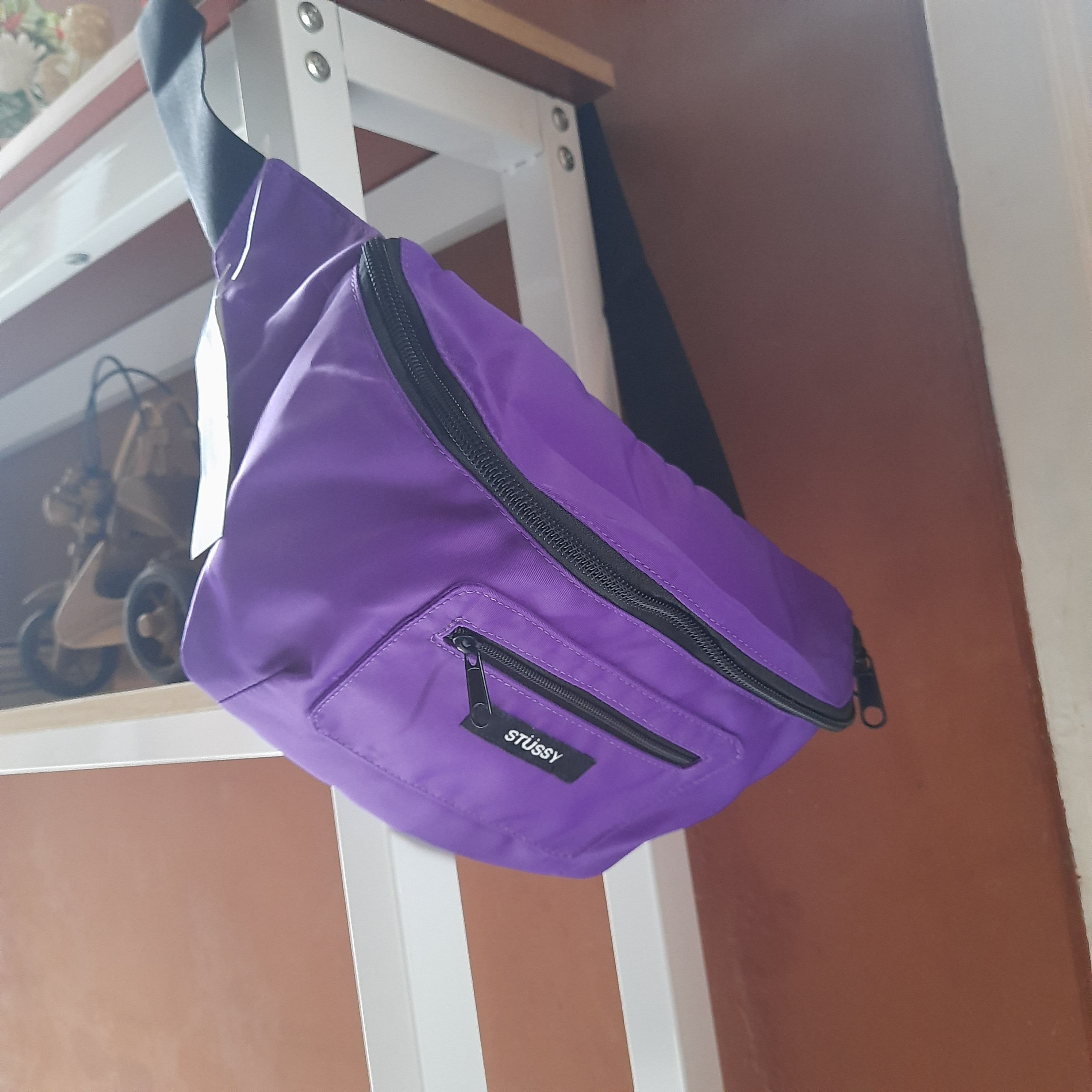 A close-up view of a purple Stussy fanny pack, casually draped over a white shelf. The pack's black zippers and Stussy logo are clearly visible against the vibrant purple fabric