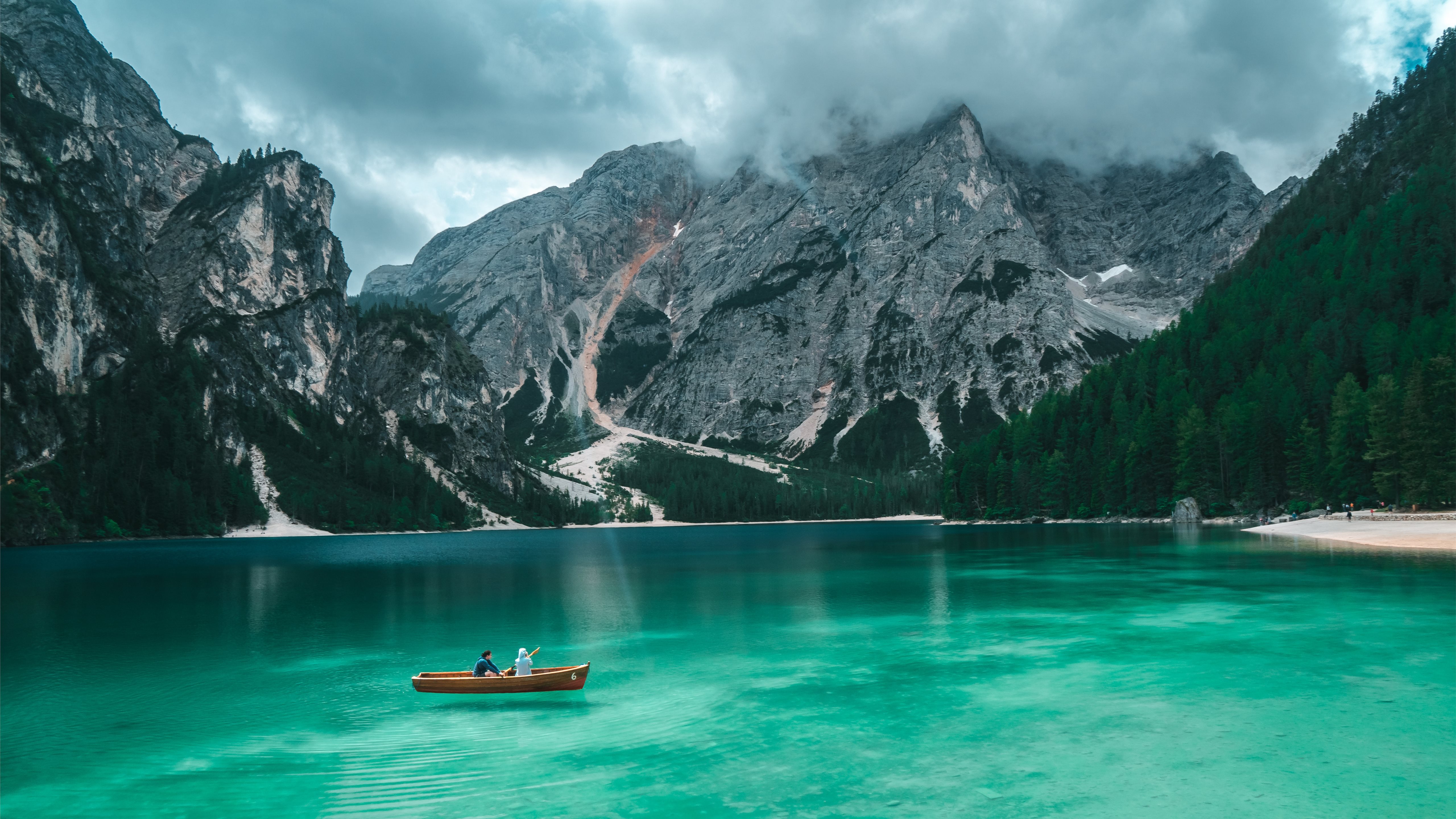 Two people in a wooden rowboat on a pristine turquoise lake, surrounded by towering, dramatic mountains and lush green forests. The cloudy sky adds to the serene yet awe-inspiring atmosphere