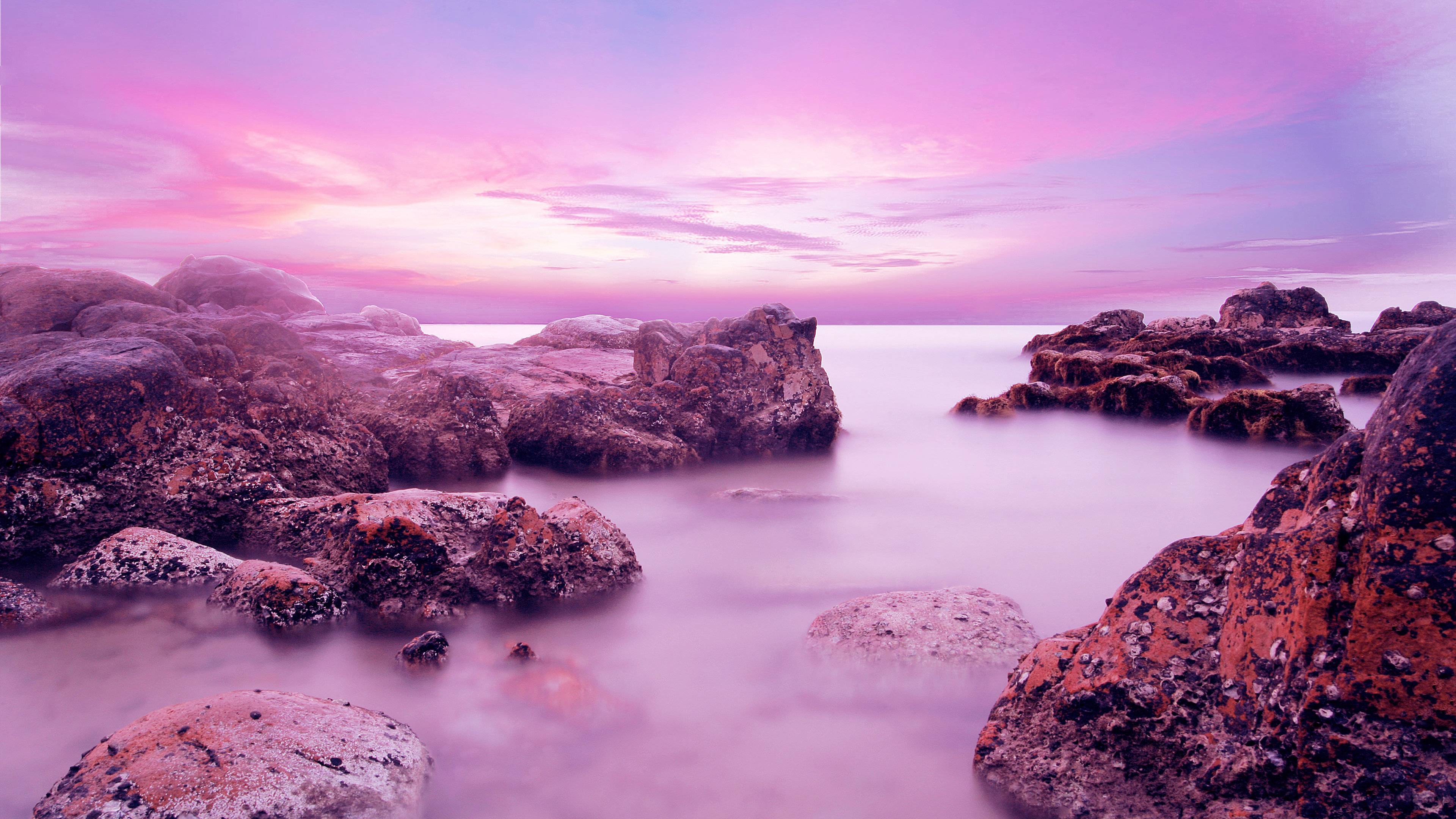 A breathtaking 4k wallpaper capturing a serene seascape at sunset. Smooth, pink-hued water flows around moss-covered rocks, creating a tranquil and dreamy atmosphere