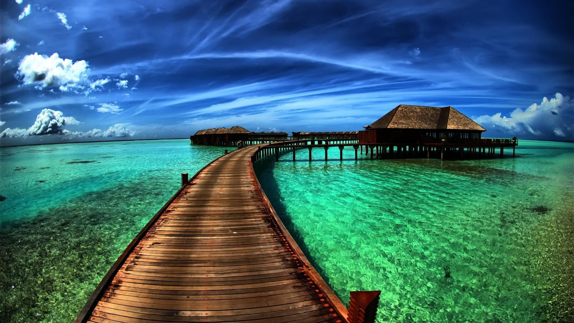 A wooden walkway leading to overwater bungalows in crystal-clear turquoise water, with a vibrant blue sky and fluffy white clouds