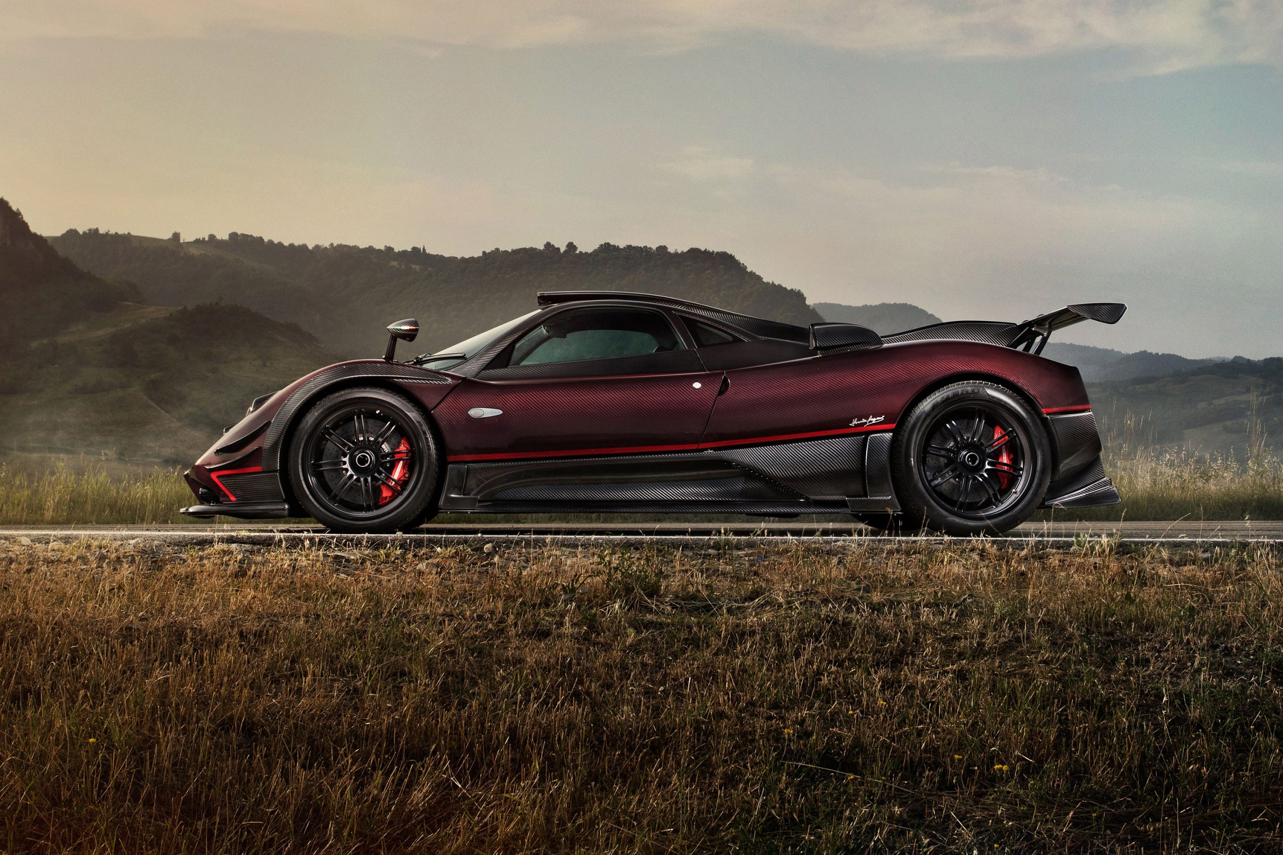 A side view of a sleek, maroon Pagani Huayra supercar parked on the roadside, set against a backdrop of rolling hills