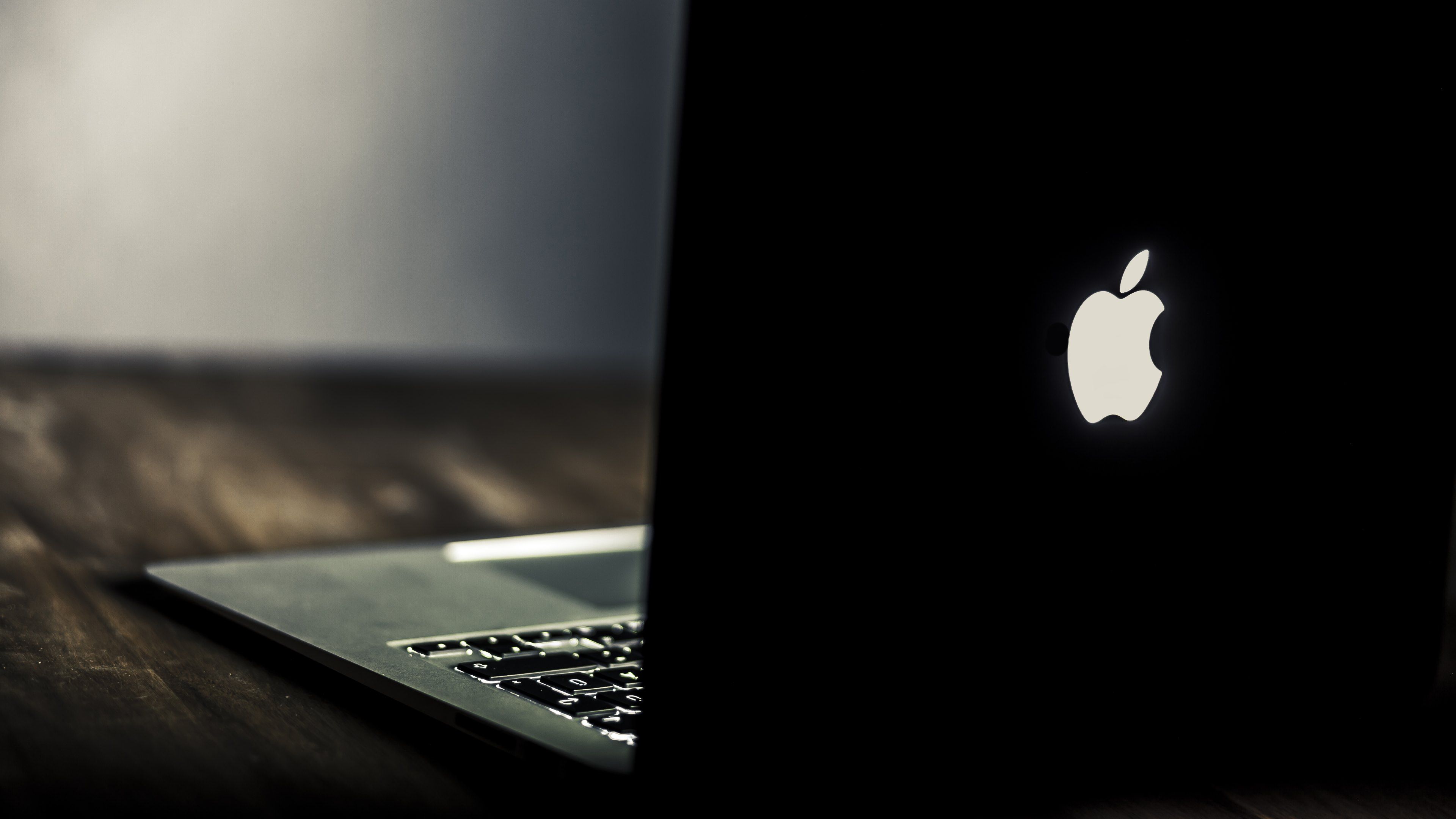 Dark image of a MacBook Pro with the Apple logo illuminated on the back, resting on a wooden table