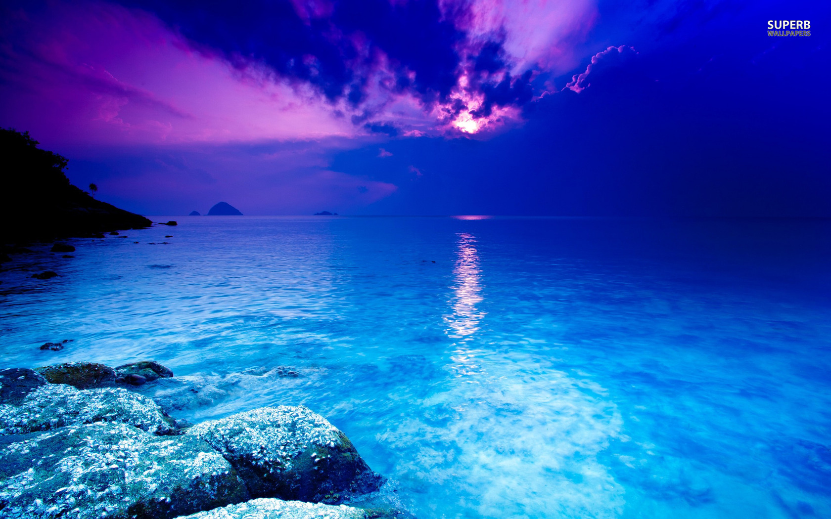 A breathtaking sunset over a tranquil turquoise sea, with the vibrant hues of purple and blue reflecting on the calm waters. Rocky foreground adds depth to the serene coastal landscape