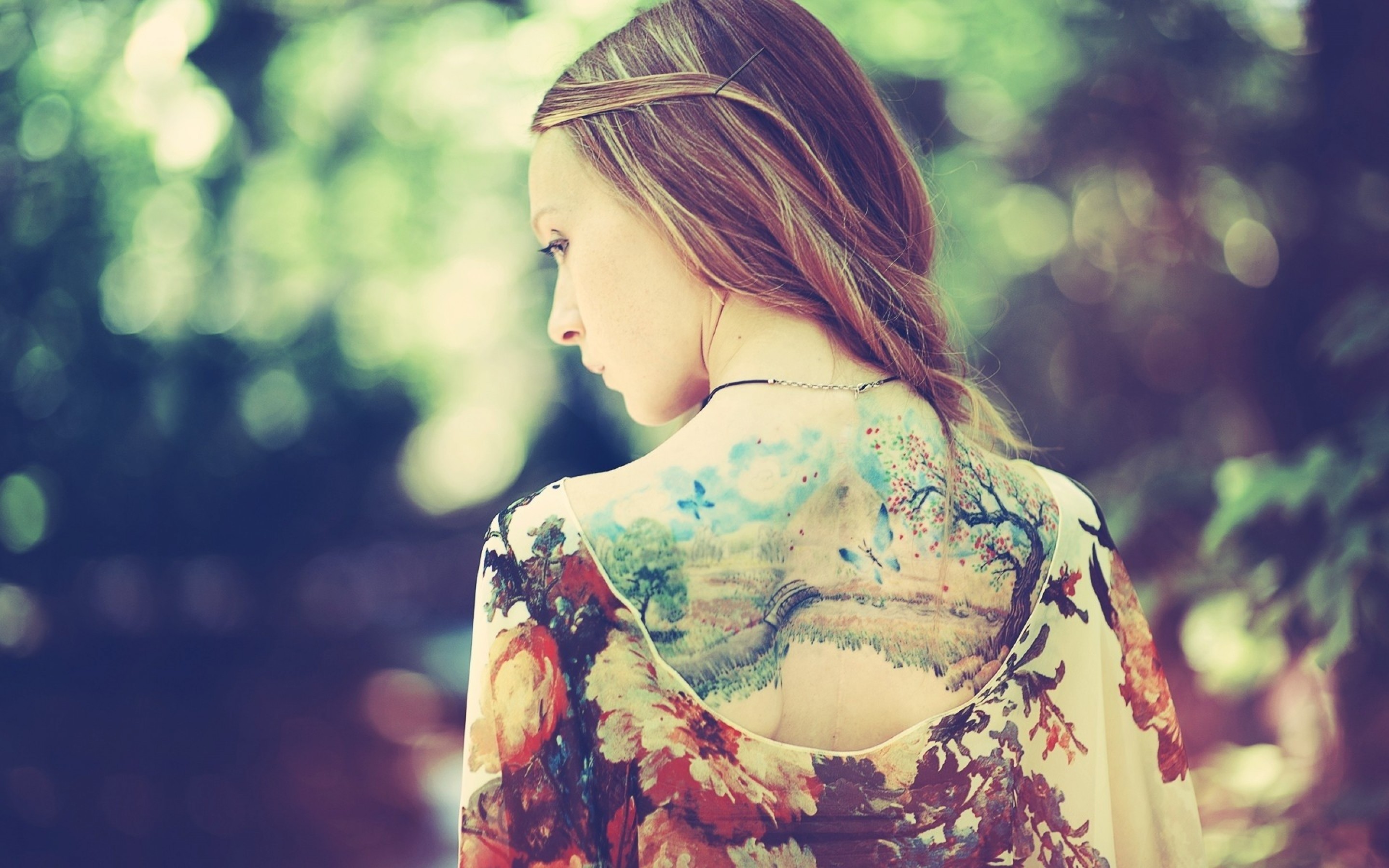 A woman with a nature-inspired tattoo on her back, featuring a landscape with trees, a bridge, and blue accents. She's wearing a floral blouse and her hair is styled in a half-updo