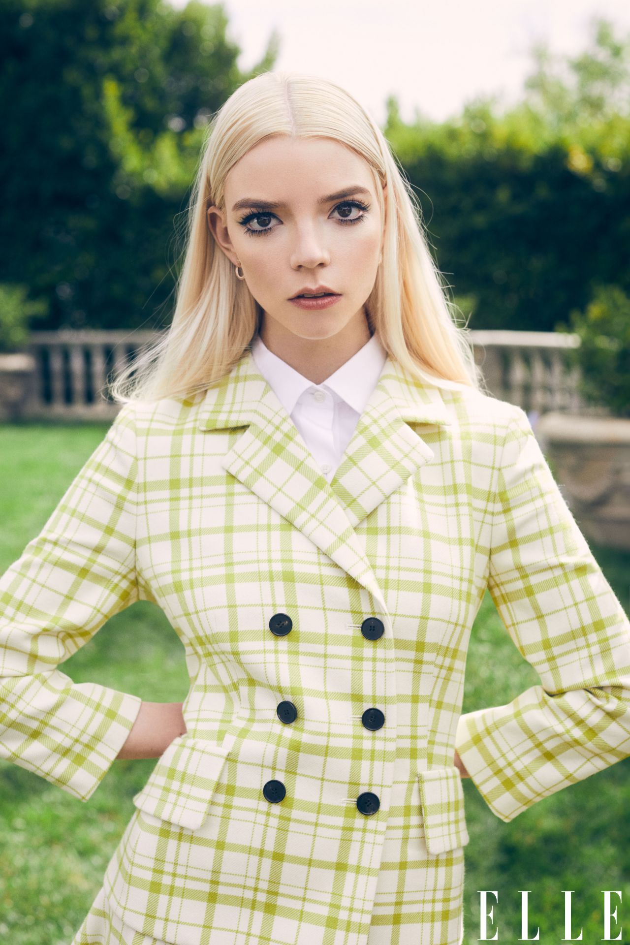 Anya Taylor-Joy posing in a green and white checkered blazer and white collared shirt in front of a green lawn