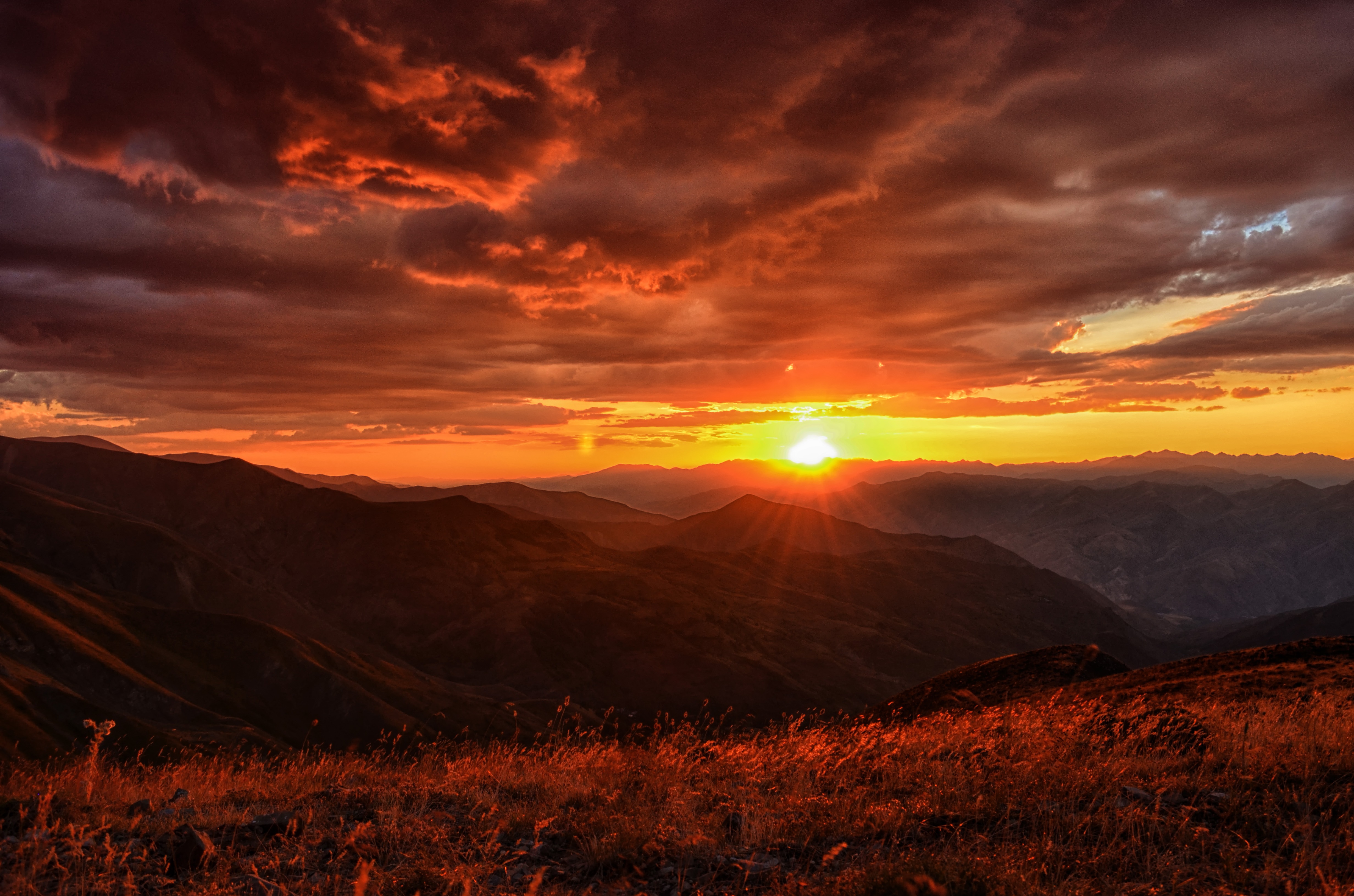 A breathtaking sunset casts a warm, orange glow over a rugged mountain range. The setting sun peeks through the clouds, creating a dramatic and vibrant scene