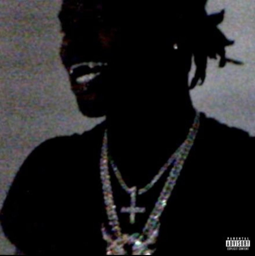 Ken Carson portrait with a slight smile, wearing multiple necklaces with a cross pendant, set against a dark background