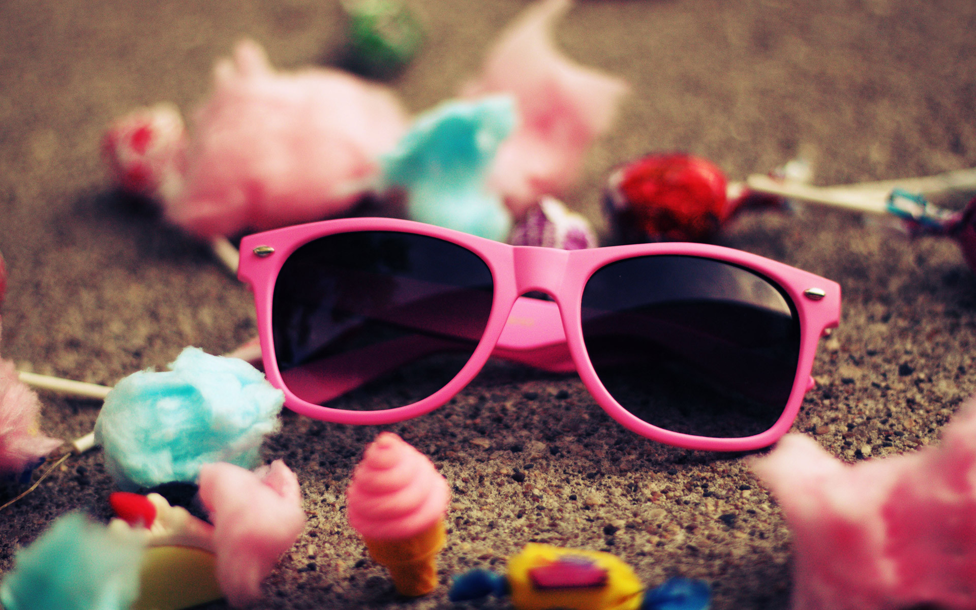 Pink sunglasses on a sandy surface surrounded by scattered candies and cotton candy