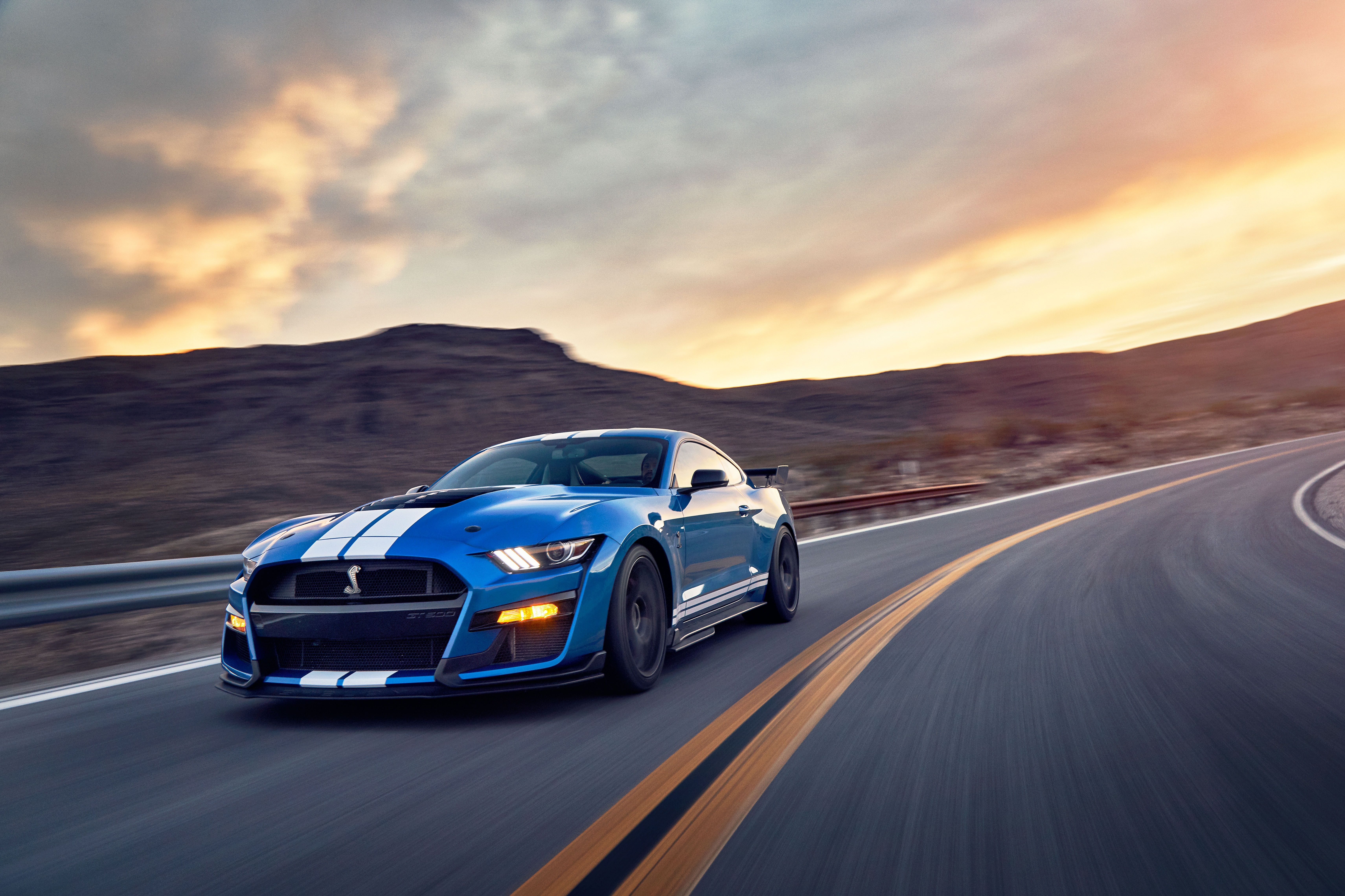 A blue Ford Mustang Shelby GT500 speeding down a desert highway at sunset