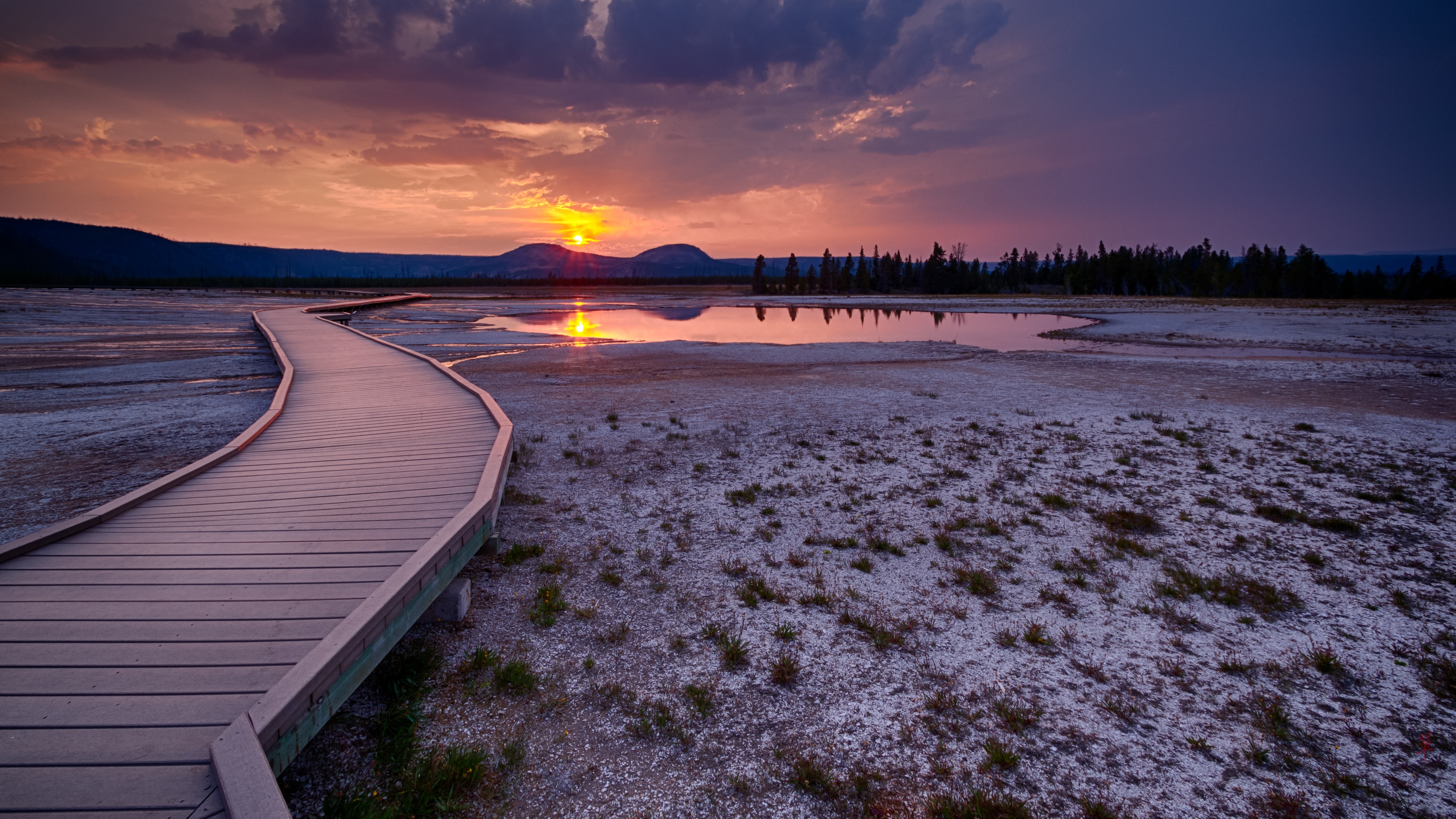 A stunning 4k background image of a wooden boardwalk winding through a geyser basin at sunset. The sky is a vibrant mix of purple and orange hues, reflecting in the still water of the geyser pool.  A distant treeline silhouettes against the colorful sky
