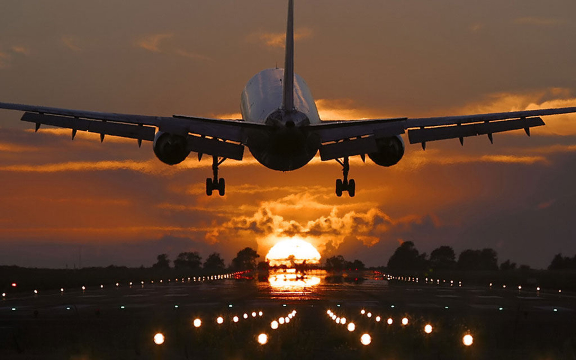 A passenger airplane silhouetted against a vibrant sunset sky as it descends towards the runway, its landing gear extended and runway lights glowing
