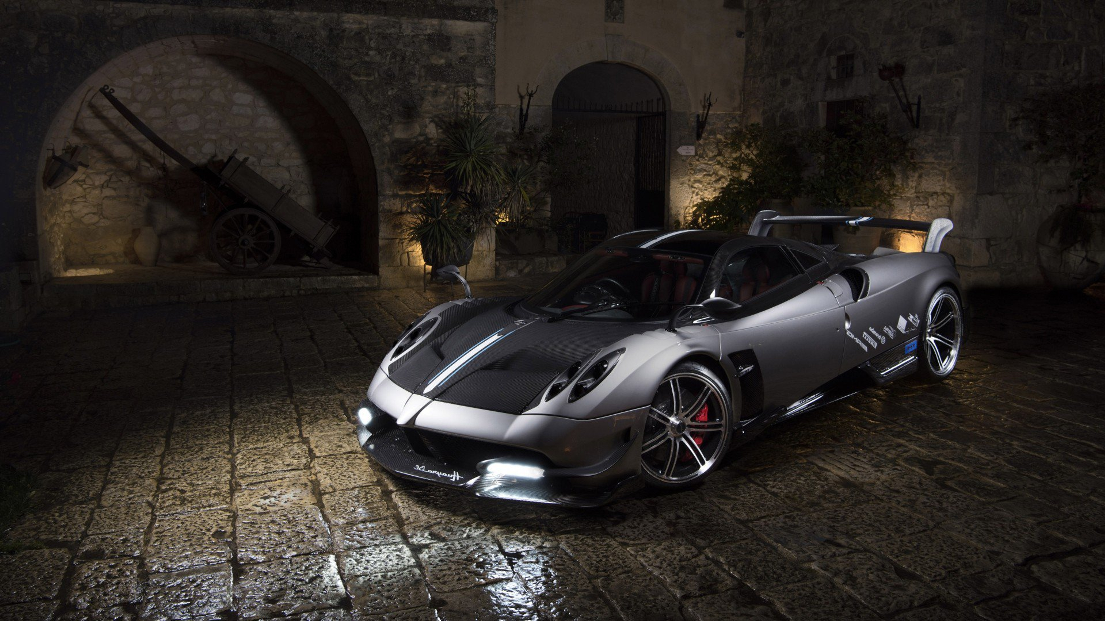 A sleek, silver Pagani Huayra supercar parked on a cobblestone street at night, with a historic building and an old cart in the background
