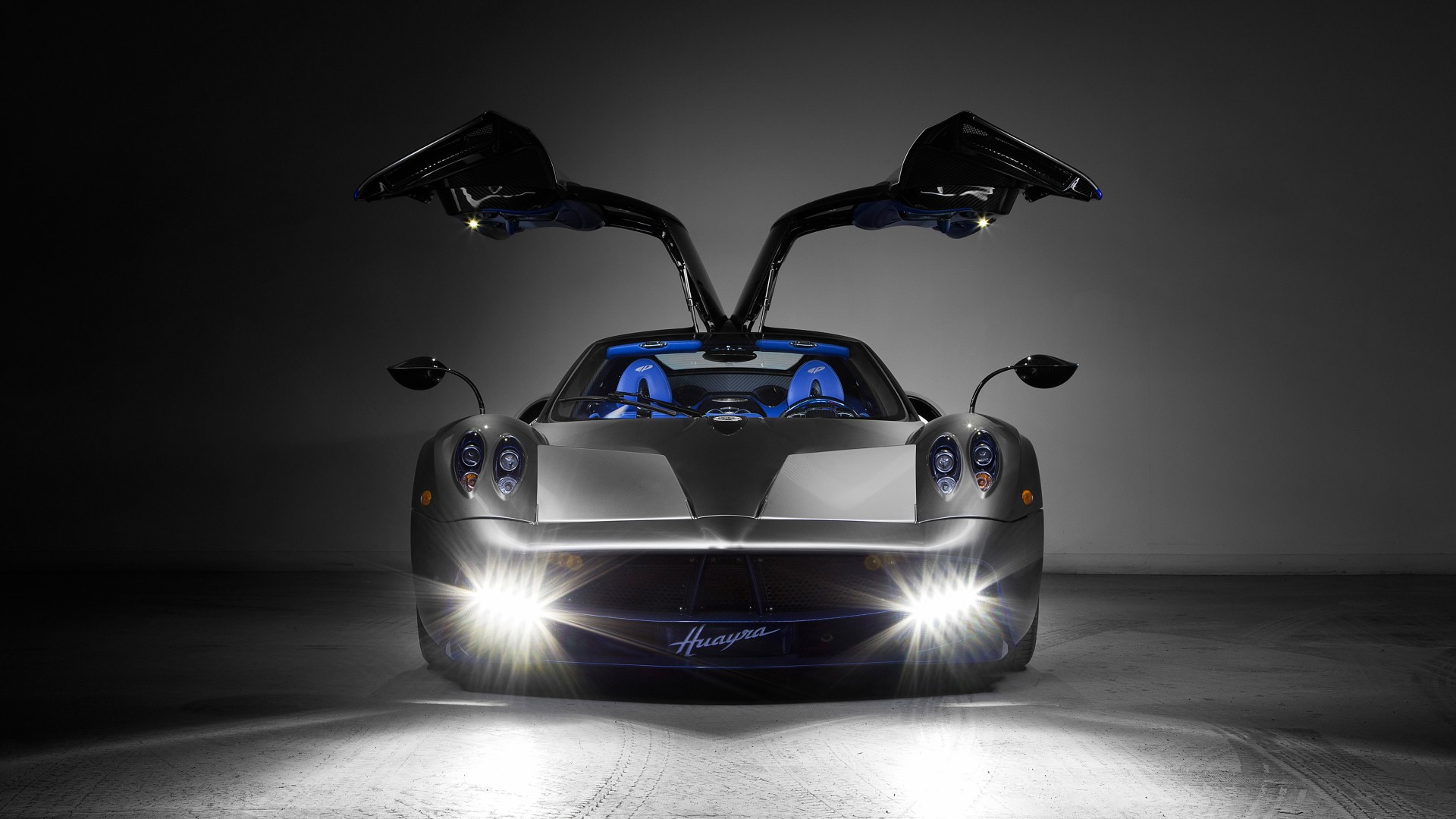 A Pagani Huayra supercar with its iconic gullwing doors open, illuminated by bright headlights in a dark setting
