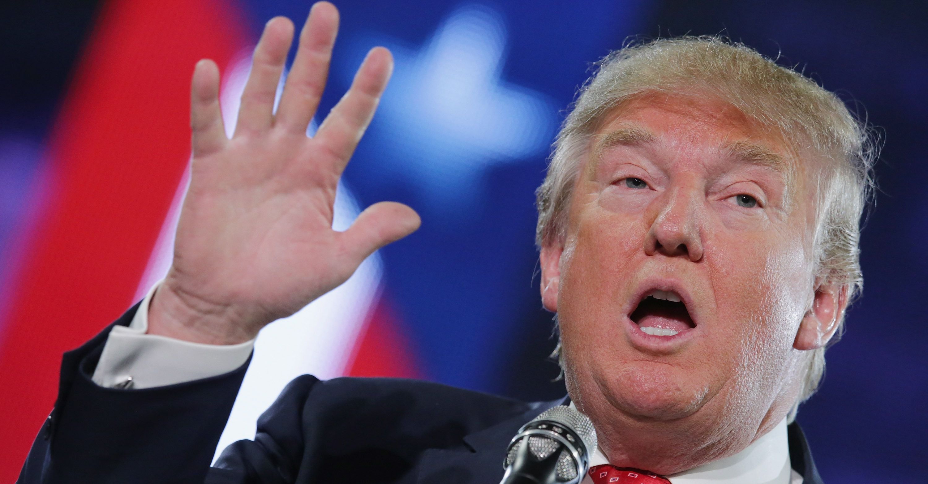 Donald Trump speaking at a rally, with a blurred American flag in the background