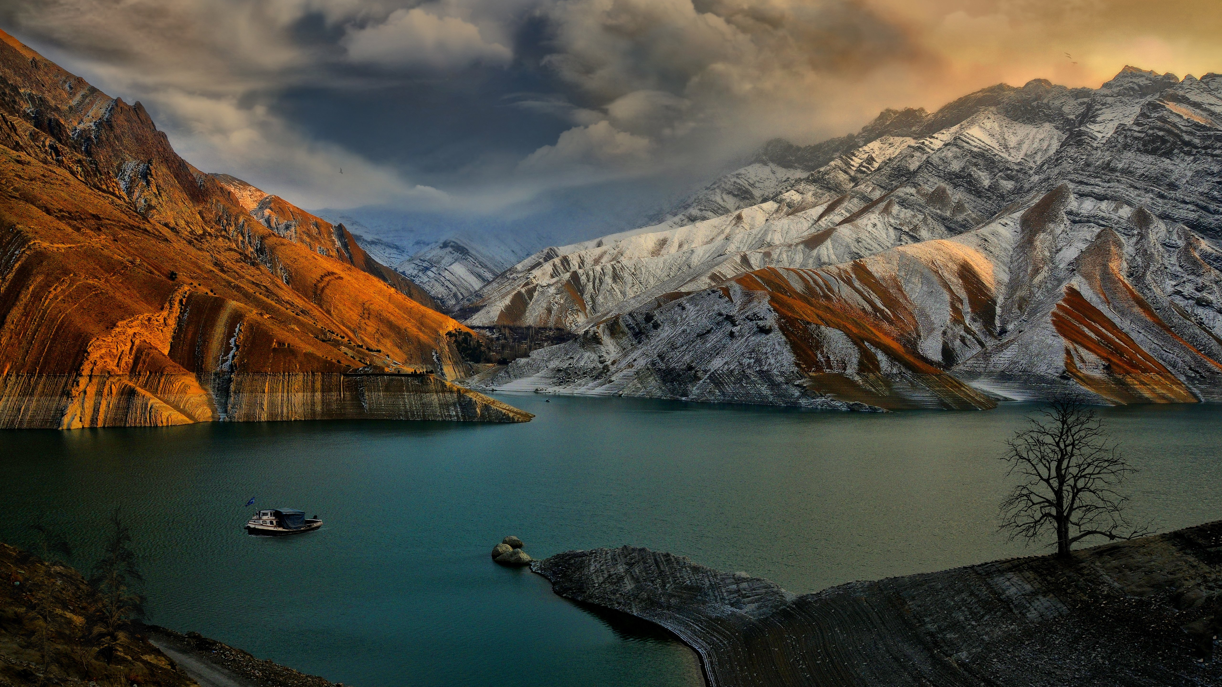 A breathtaking 5K wallpaper showcasing a serene lake nestled amidst snow-capped mountains, with a lone boat and tree adding to the picturesque scene