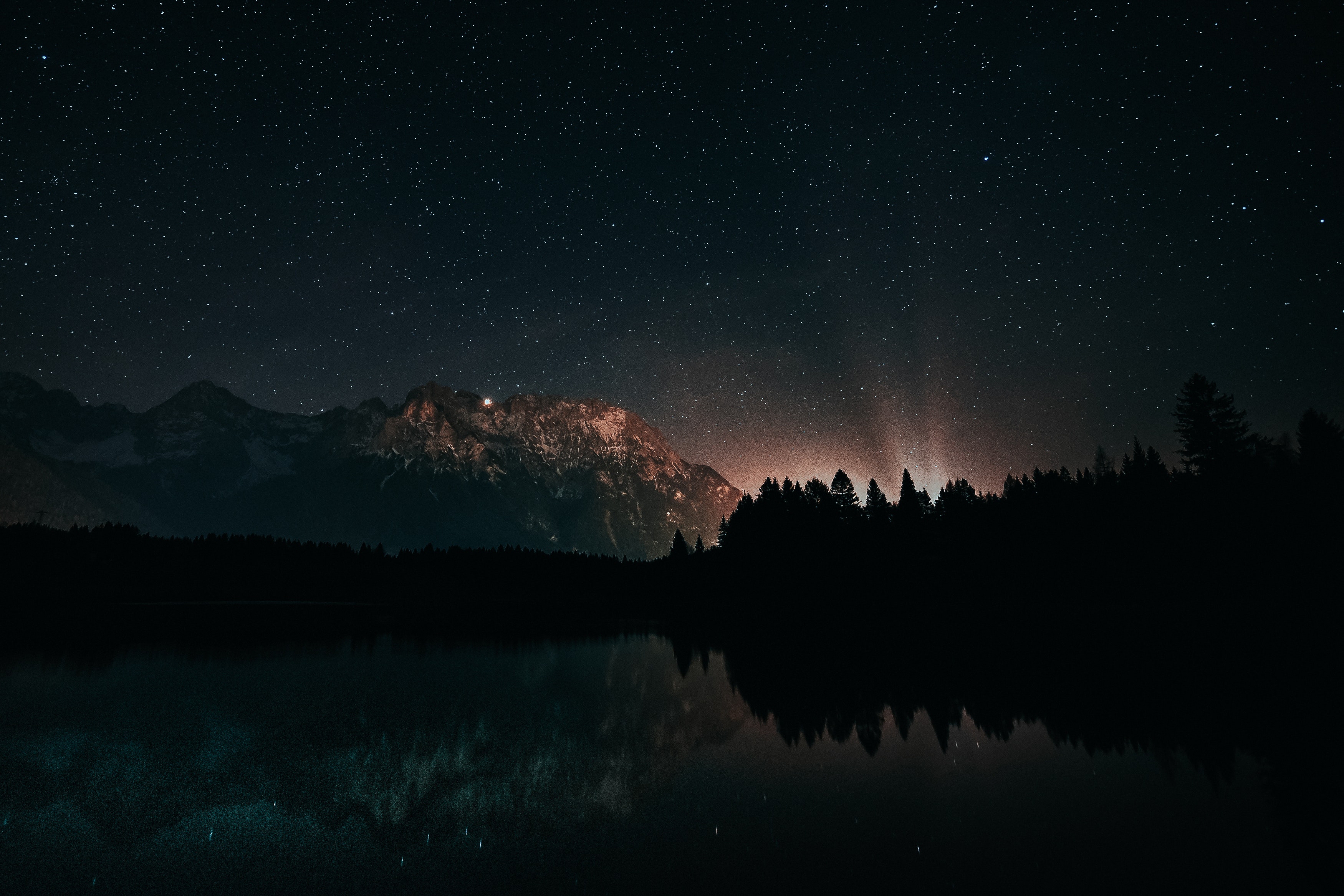 A breathtaking astrophotography image capturing a serene lake reflecting the starry night sky and a silhouetted mountain range. The Milky Way galaxy is faintly visible, adding to the magical ambiance