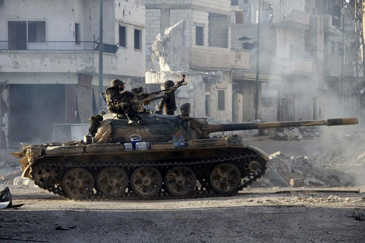 A Syrian tank advances through the rubble-strewn streets of a war-torn city in Syria. Two soldiers man the tank, one aiming a machine gun