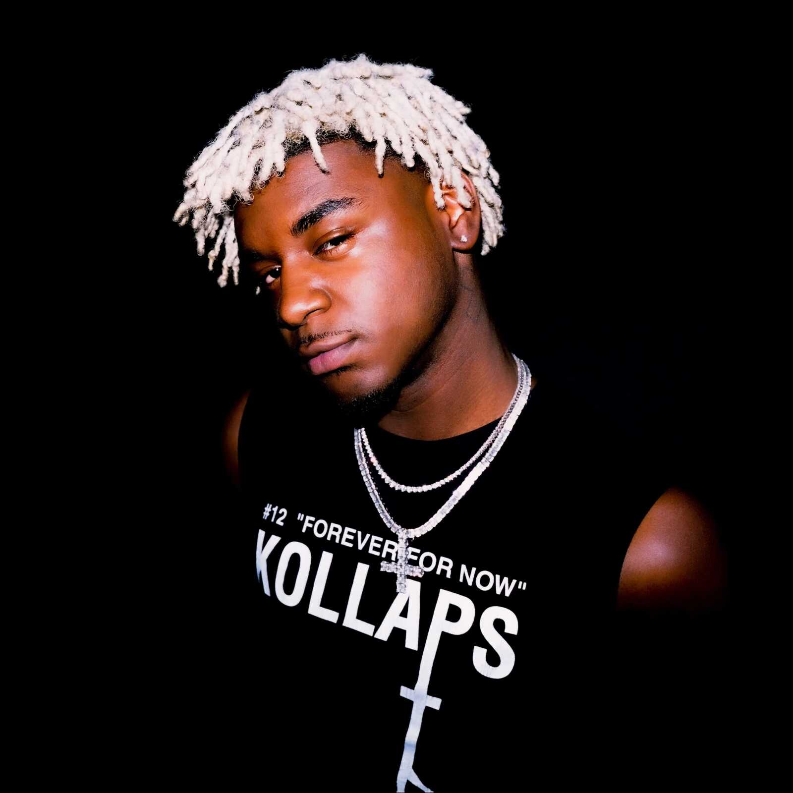 Ken Carson posing in a black sleeveless shirt with his signature bleached dreadlocks and diamond necklaces