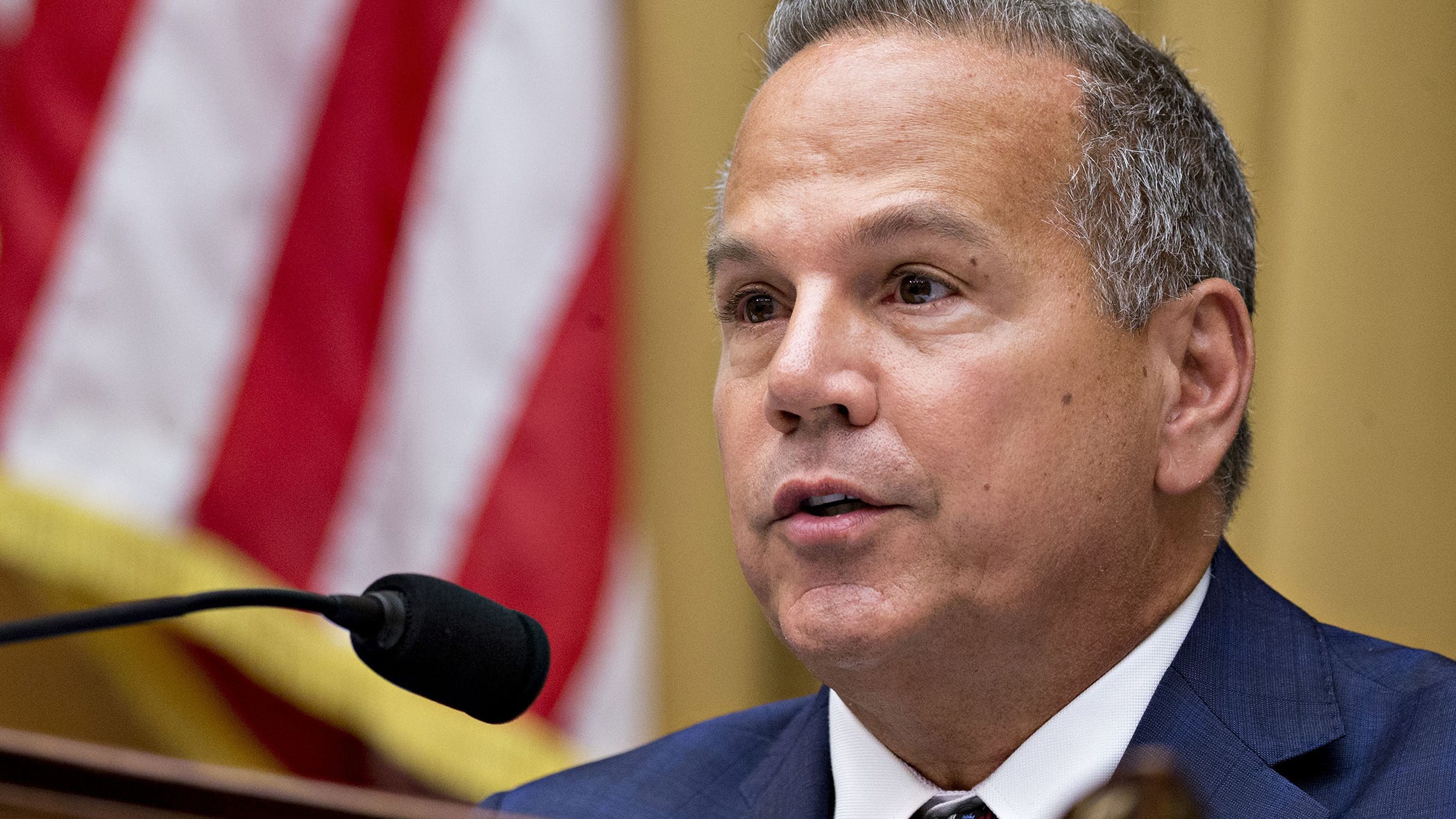 A man in a suit speaking into a microphone with an American flag in the background