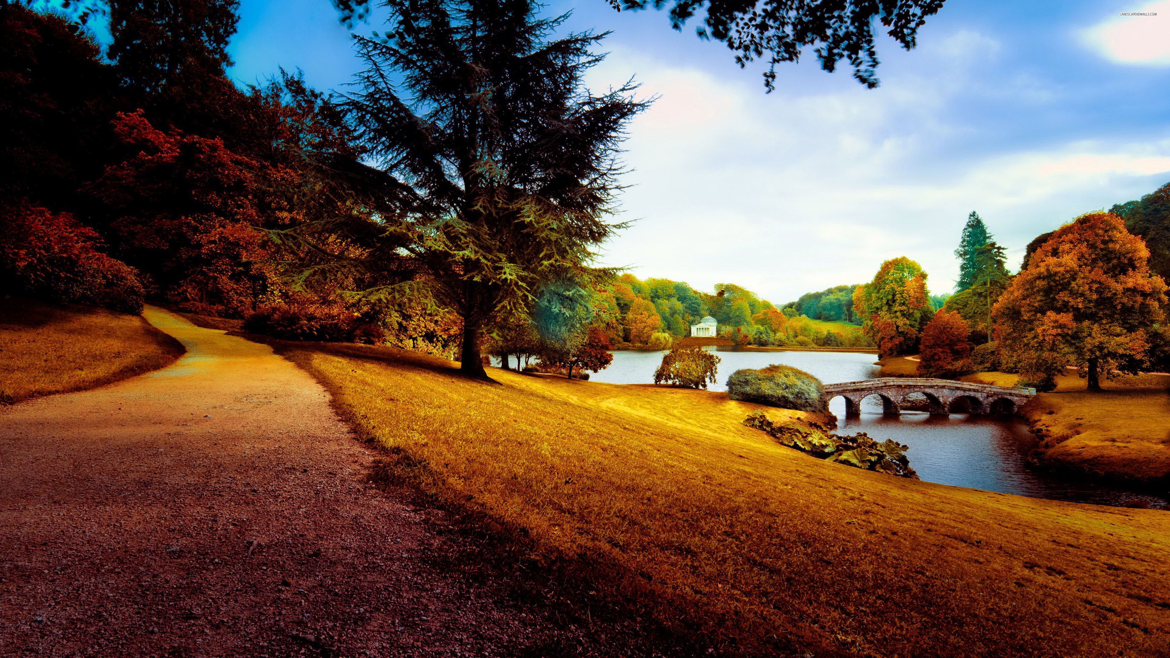 A scenic 4K wallpaper featuring a pathway meandering through an autumnal park, leading towards a stone bridge over a tranquil lake. A classical pavilion sits serenely on the far bank, framed by vibrant fall foliage