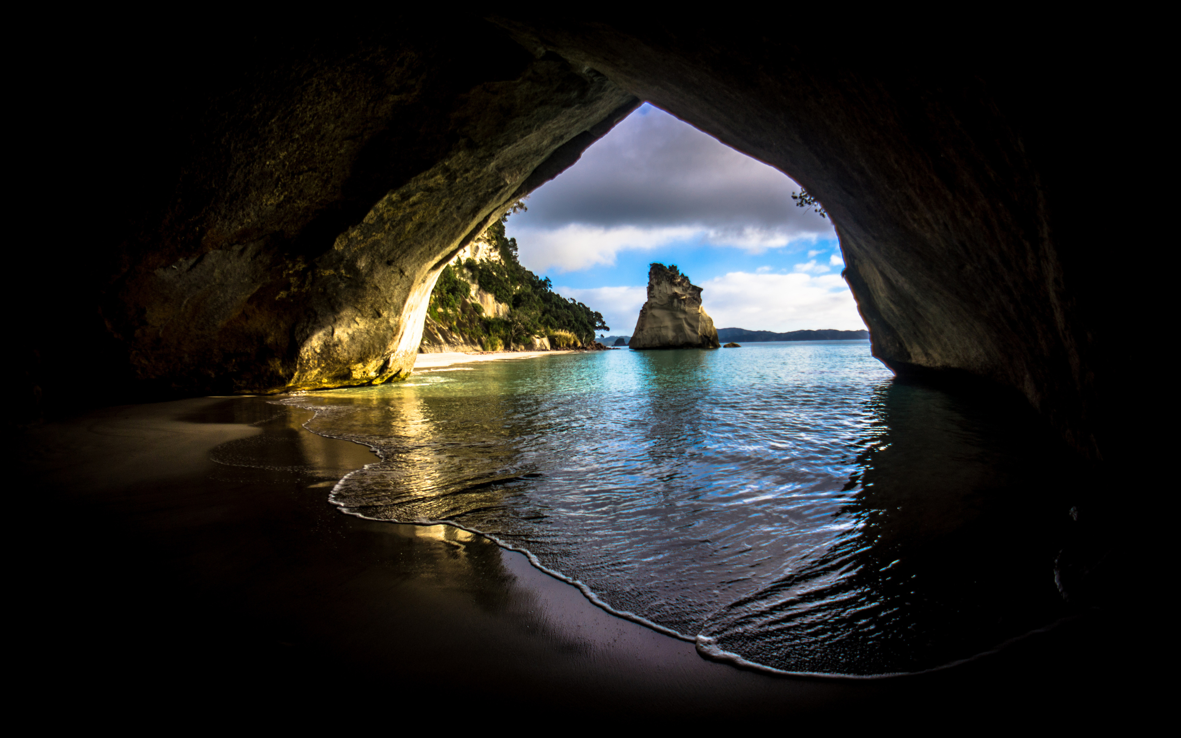 A breathtaking view from inside a dark sea cave, overlooking a tranquil beach with a unique rock formation in the turquoise water under a partially cloudy sky
