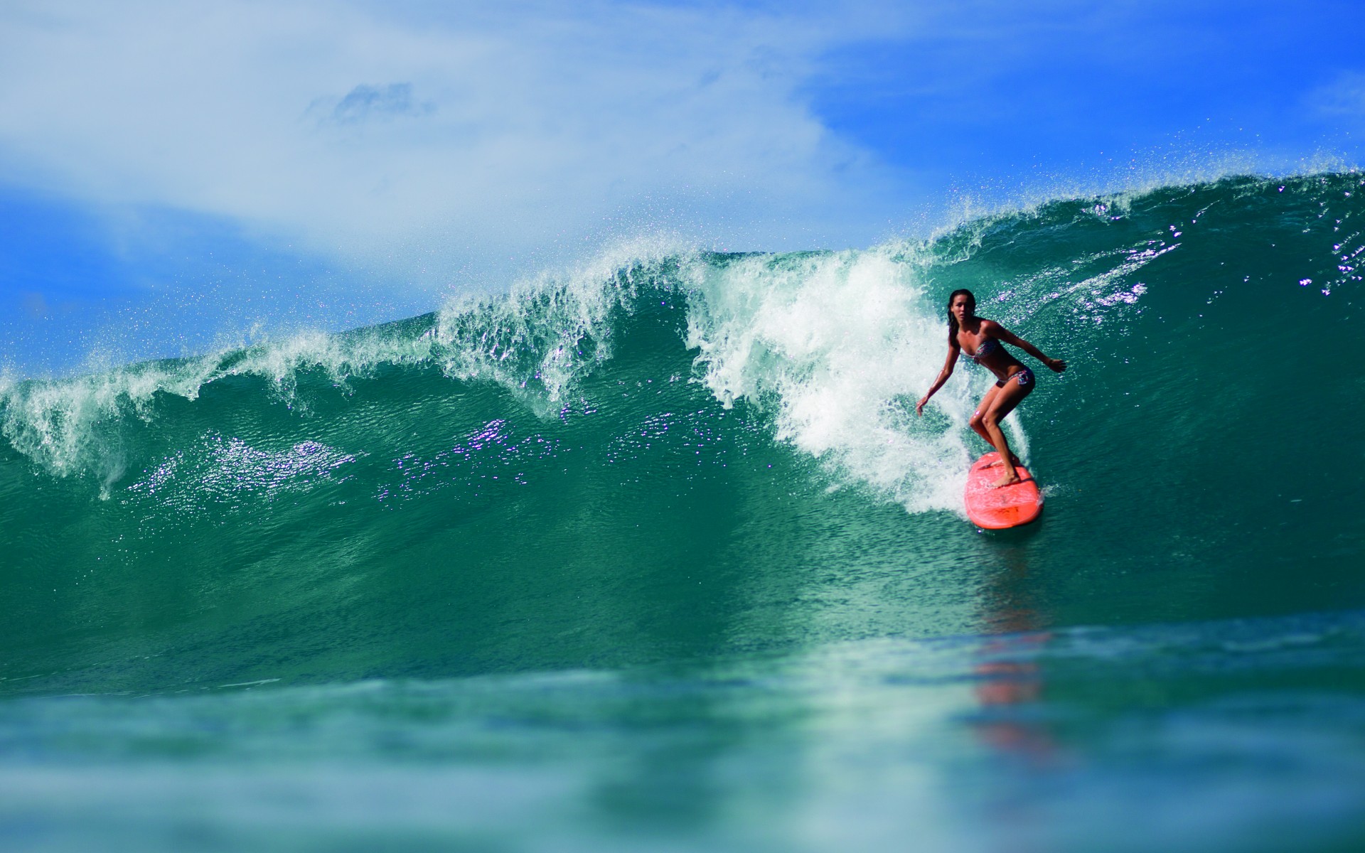 A woman surfer rides a bright orange surfboard across a vibrant turquoise wave, her form balanced and poised against the powerful curl of the ocean