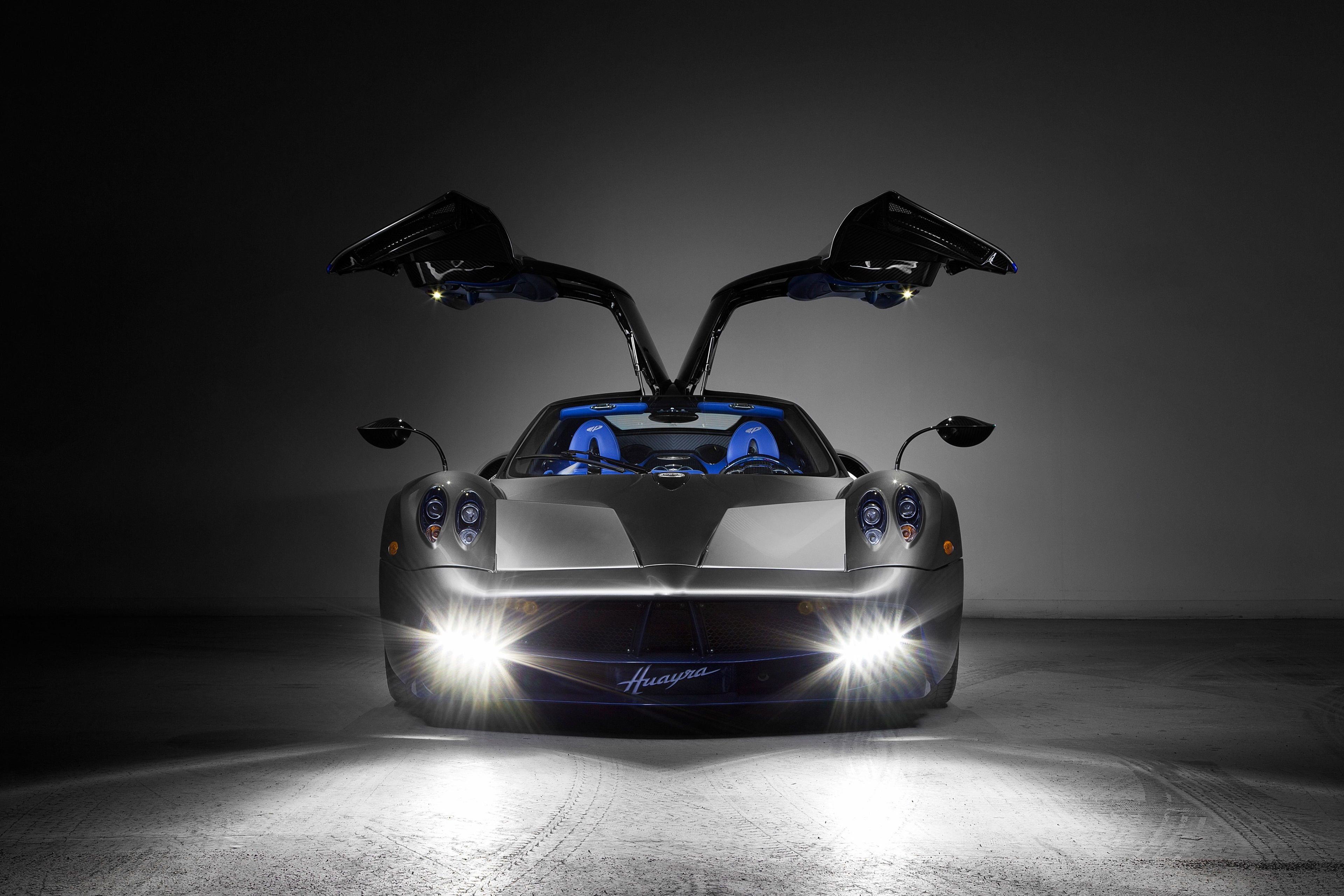 A Pagani Huayra supercar with its iconic dihedral doors open, illuminated by bright headlights in a dark setting