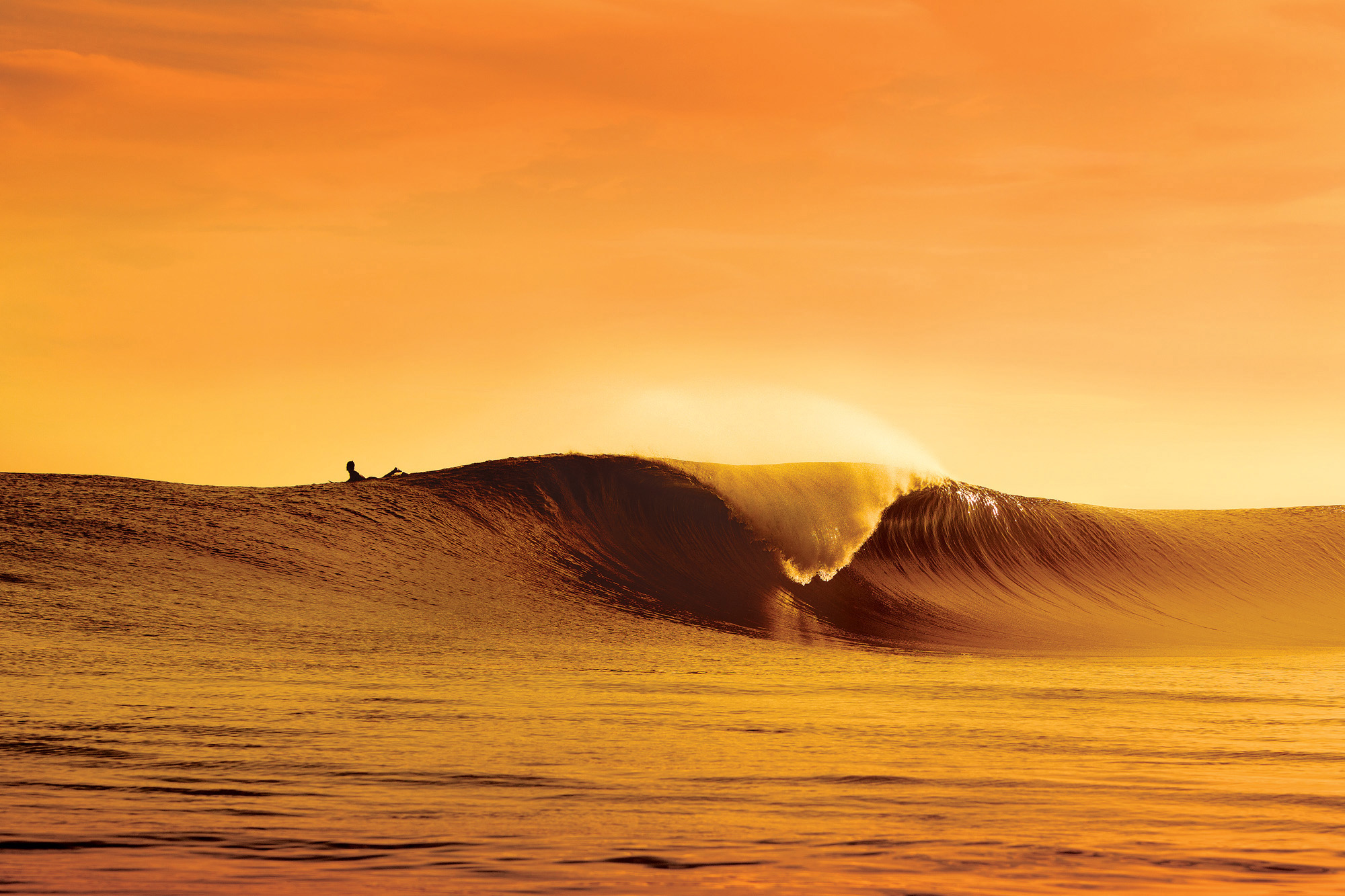 A surfer waits for the perfect wave at sunset, silhouetted against the golden sky. The wave curls invitingly, promising an exhilarating ride