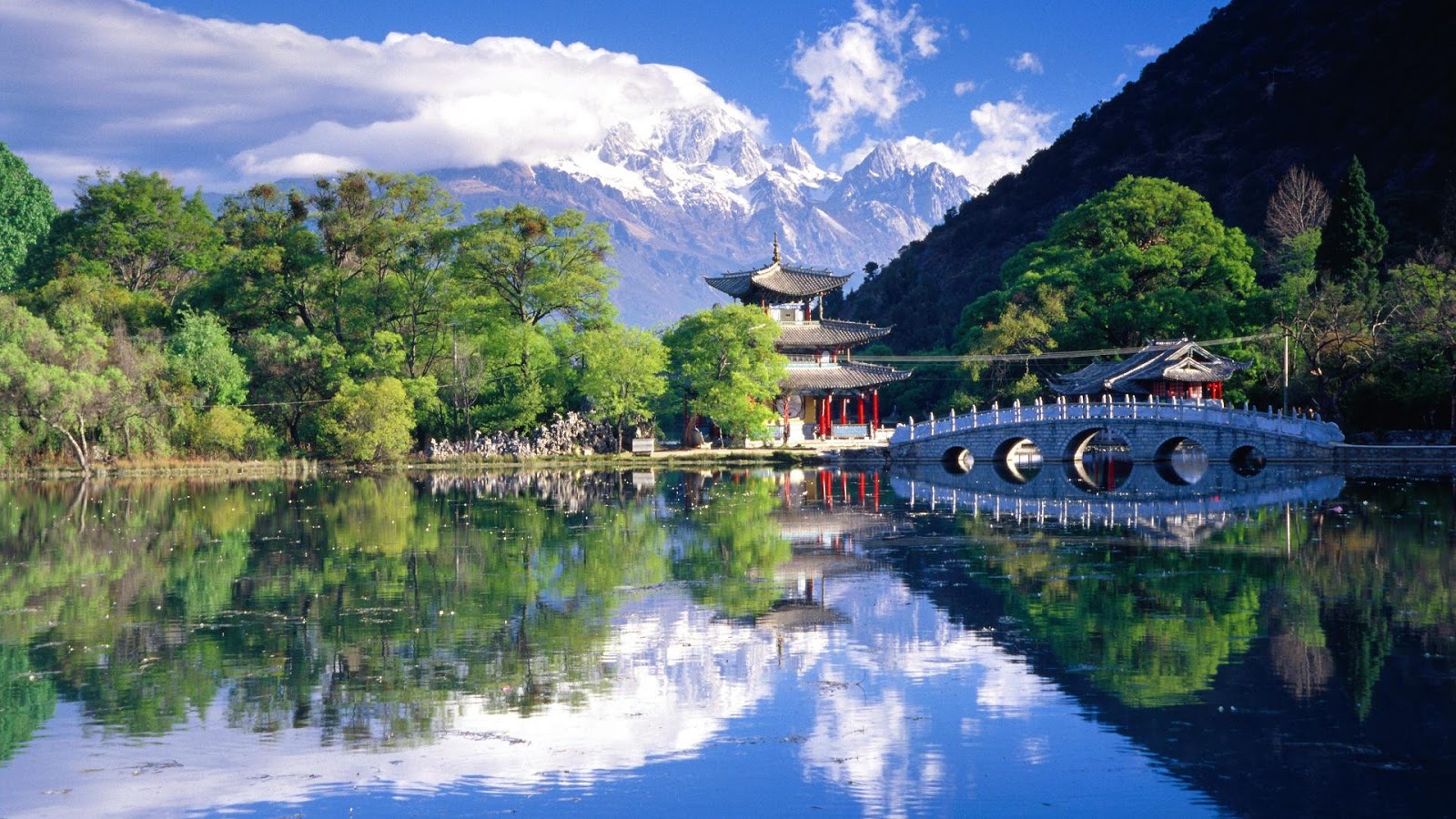A serene landscape of Black Dragon Pool in Lijiang, China, showcasing the Jade Dragon Snow Mountain, traditional Chinese architecture, and a picturesque bridge