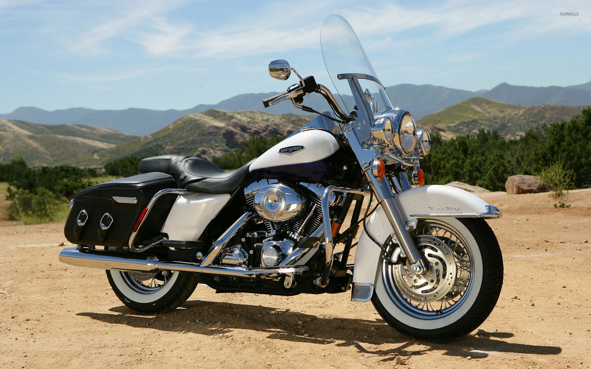 A white and chrome Harley Davidson Road King motorcycle parked on a dirt road with mountains in the background