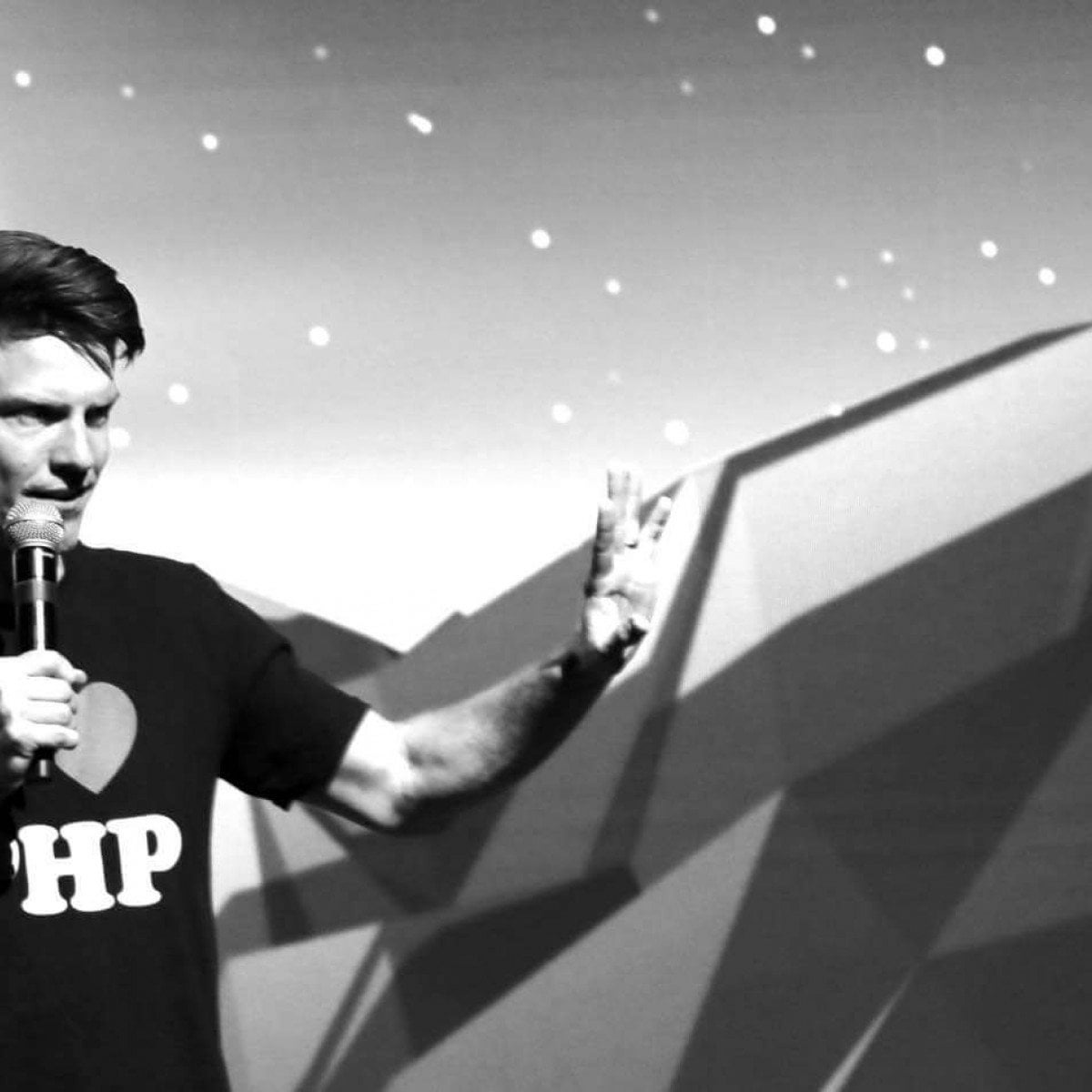 A man speaking on stage with a microphone, wearing a t-shirt with a heart and 