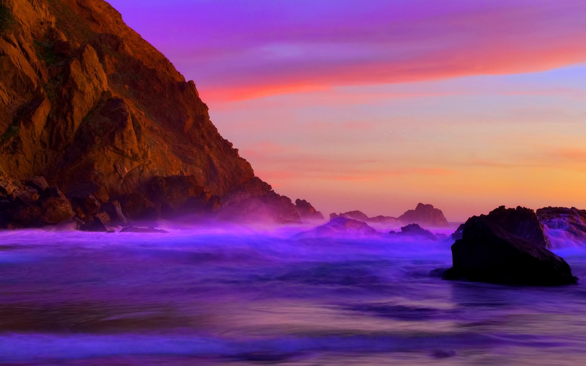 Vivid purple ocean waves crash against dark rocks at sunset, with a large cliff face in the background