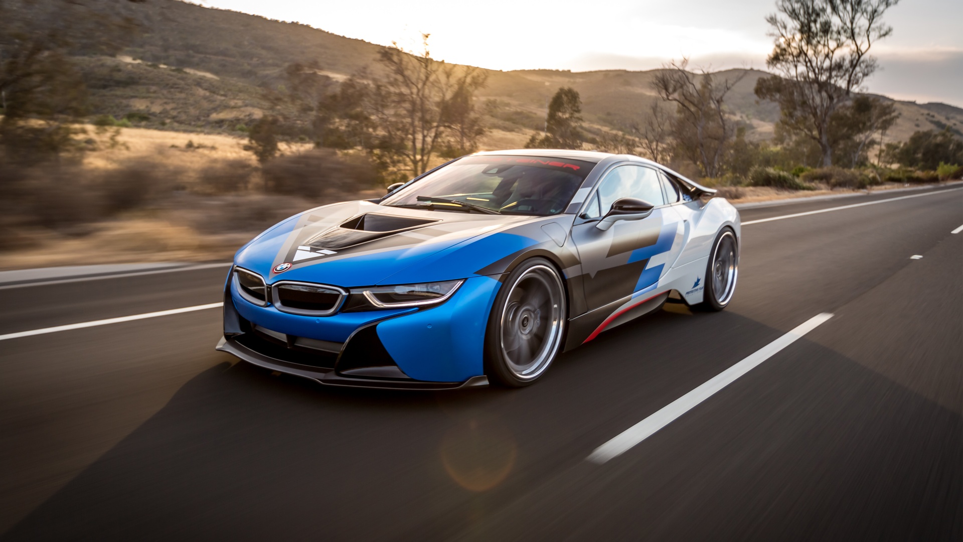 A customized BMW i8 sports car in blue and black, driving fast on a highway with a blurred background of hills and sunset