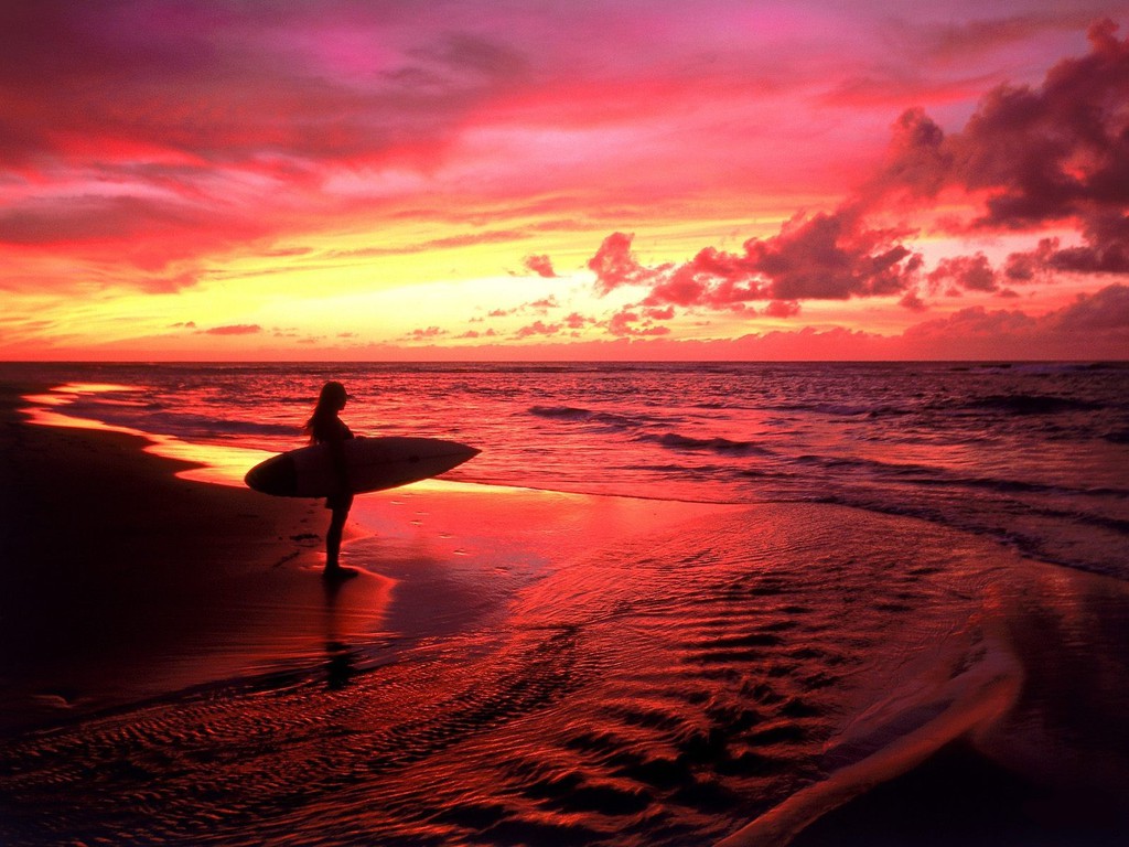 Silhouette of a surfer standing on a beach at sunset with a surfboard under their arm, the sky and water reflecting the vibrant colors of the setting sun