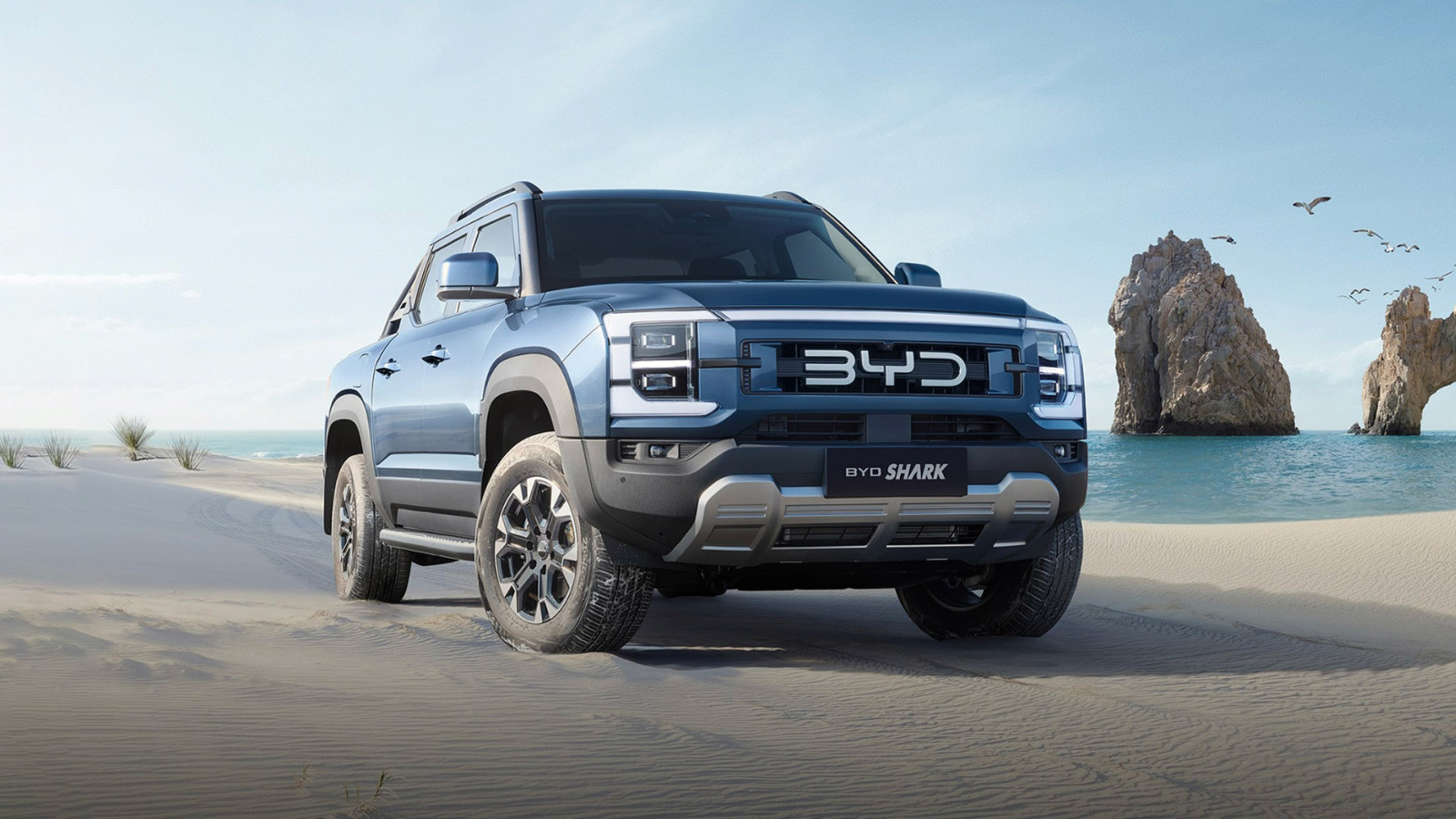 A blue BYD Shark pickup truck parked on a sandy beach, with rocky cliffs and ocean in the background