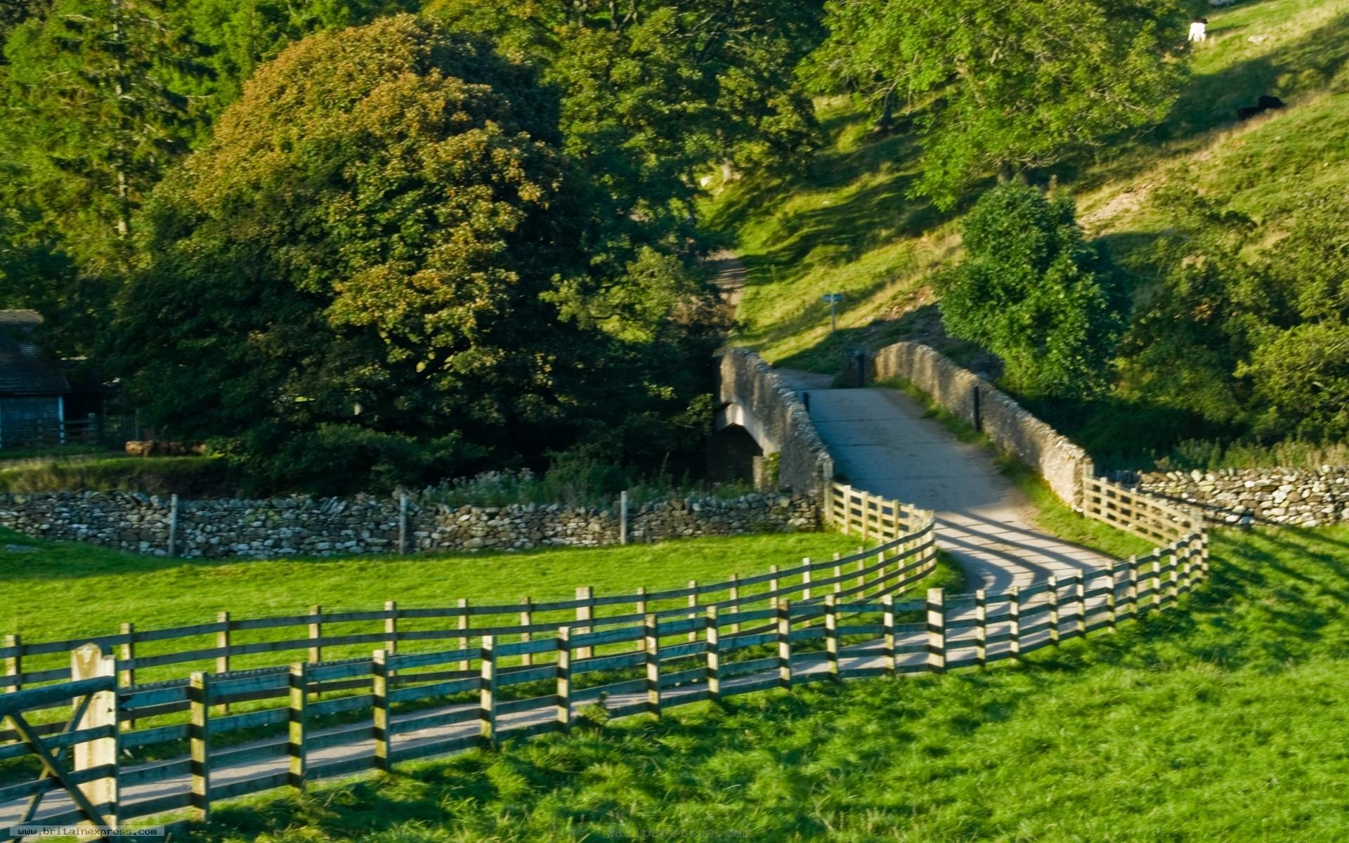 A picturesque countryside scene with a stone bridge, wooden fences, and lush green fields
