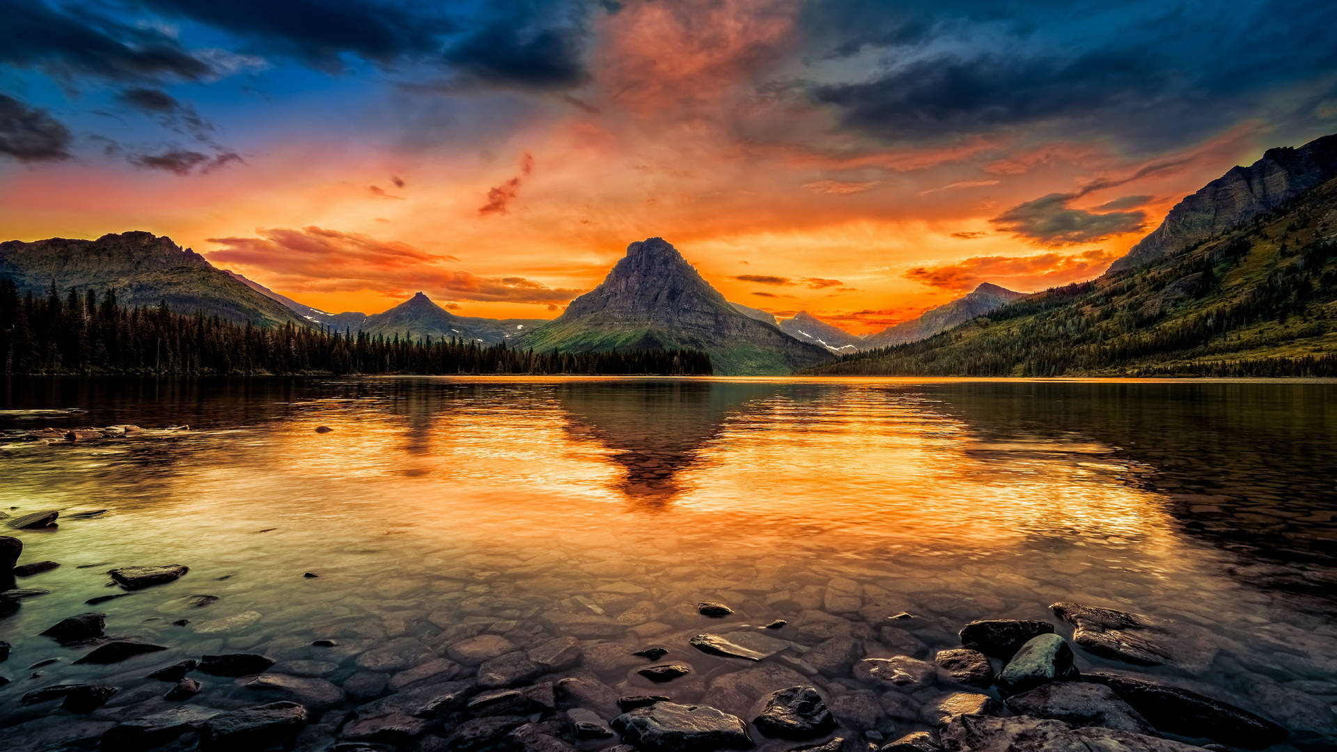 Stunning 8K HD wallpaper of a vibrant sunset over a serene mountain lake. The fiery hues of the sky are reflected in the calm water, creating a breathtaking mirror image.  Rocky shoreline details add depth and texture to this picturesque landscape