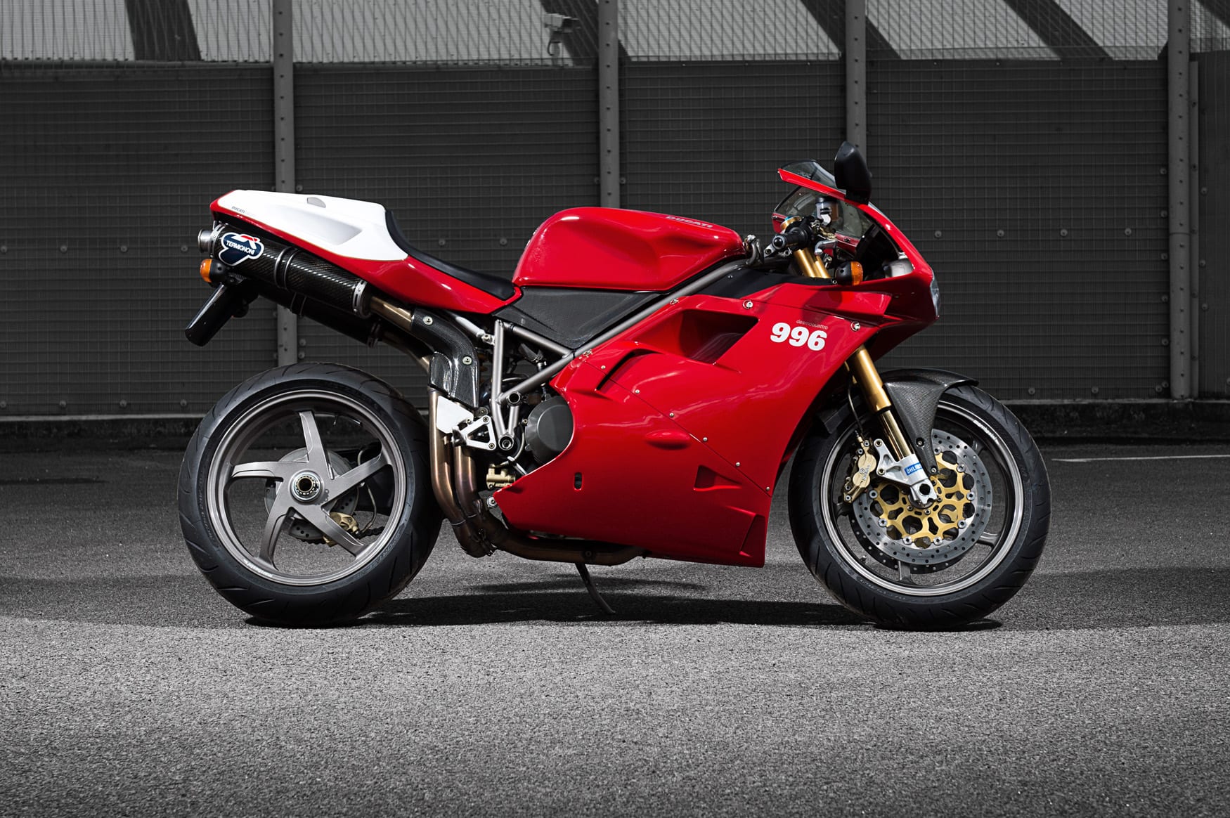 A Ducati 996 motorcycle parked in front of a metal fence.  The bike has a distinctive red body with a white tail section