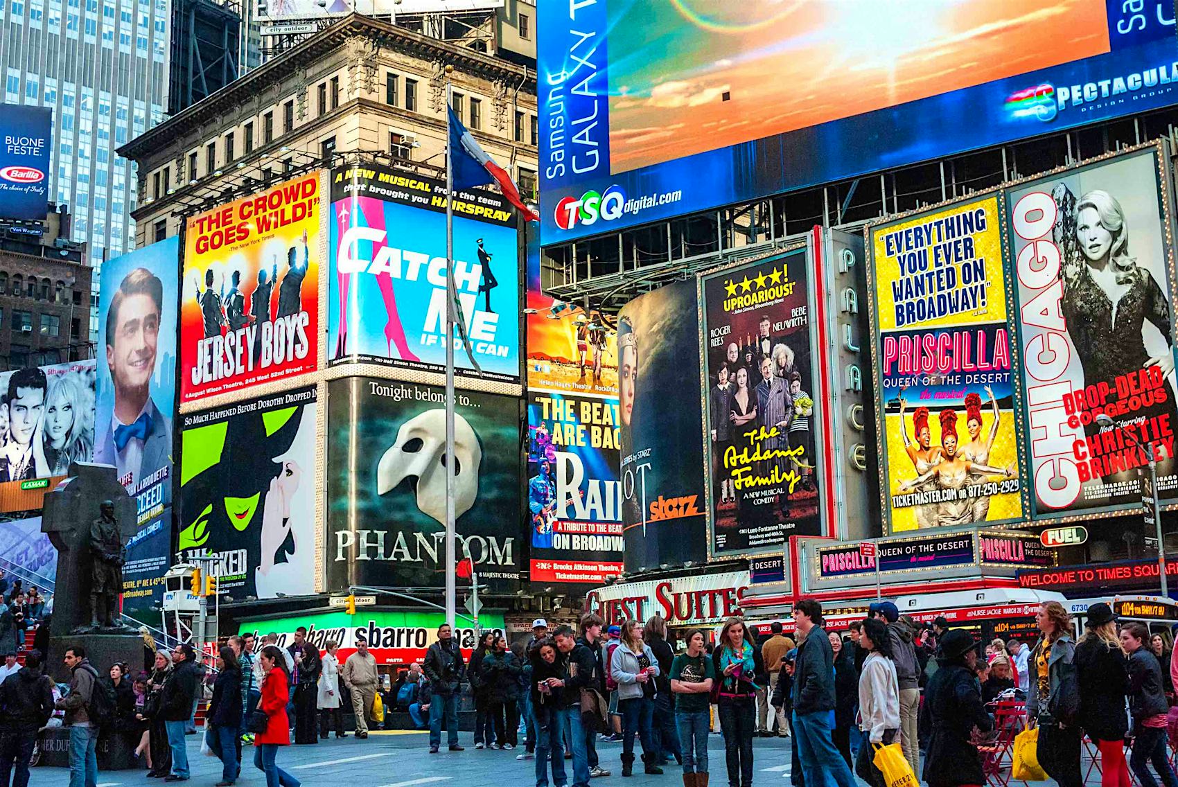 A vibrant view of Times Square in New York City at night, showcasing numerous brightly lit billboards advertising Broadway shows like Jersey Boys, Catch Me If You Can, Phantom of the Opera, The Addams Family, Priscilla Queen of the Desert, and Chicago. The bustling square is filled with people enjoying the lively atmosphere