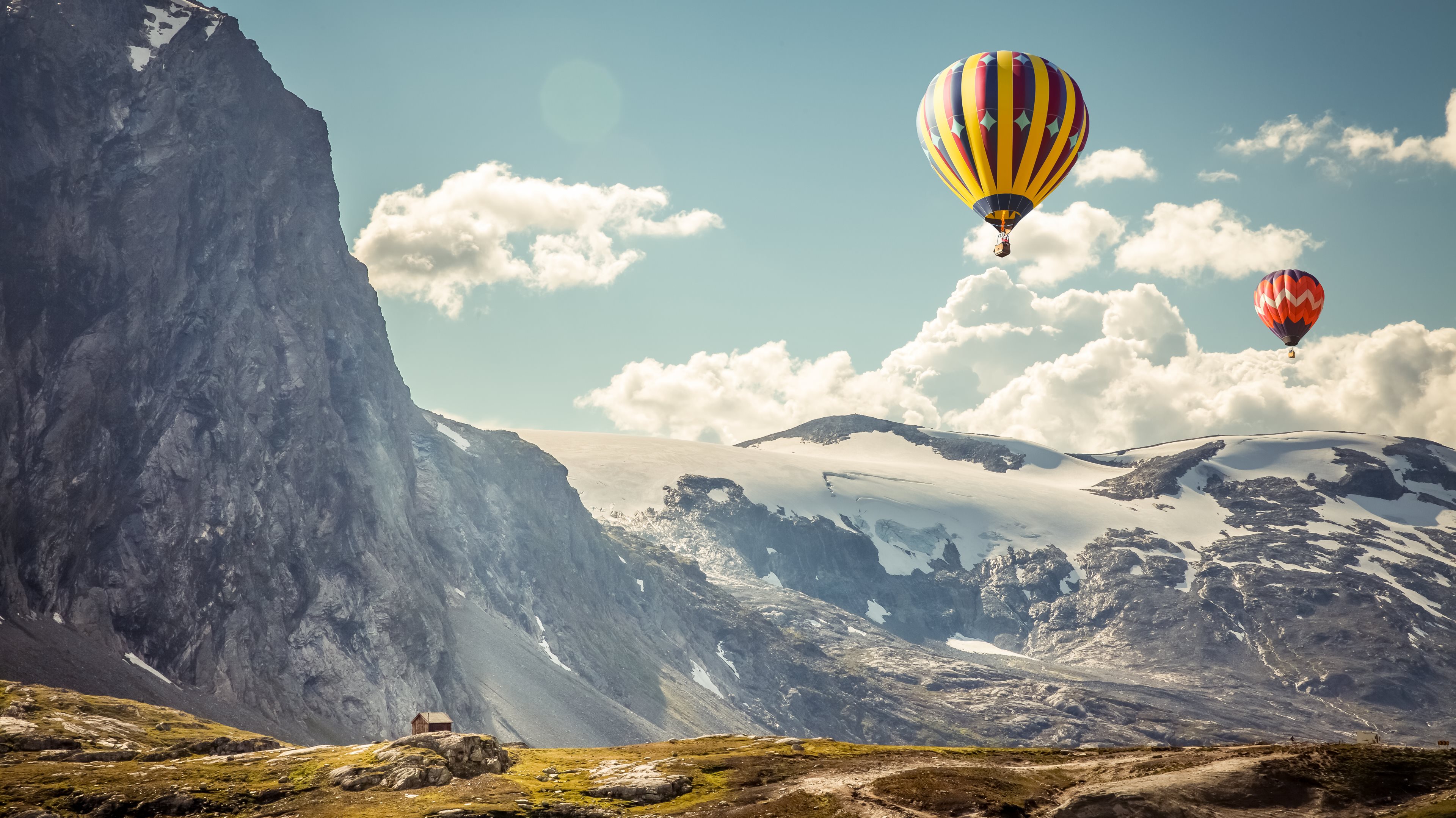 Two hot air balloons soar over a breathtaking mountain landscape with snowy peaks and a small cabin nestled below