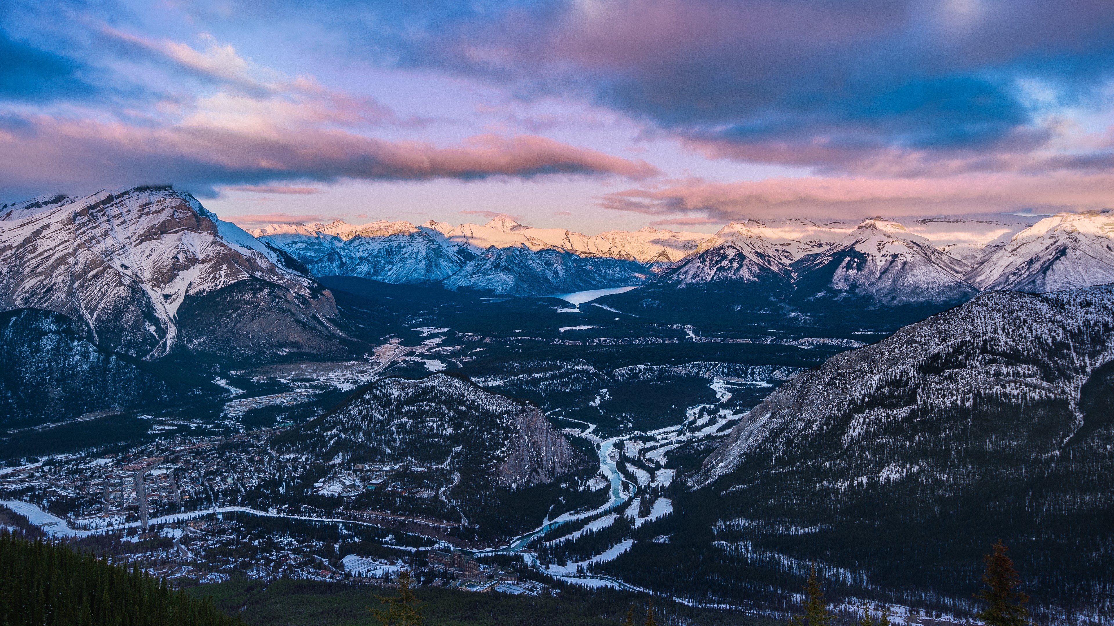 A breathtaking panoramic view of snow-capped mountains at sunset, featuring a valley with a winding river and a picturesque town in the Canadian Rockies