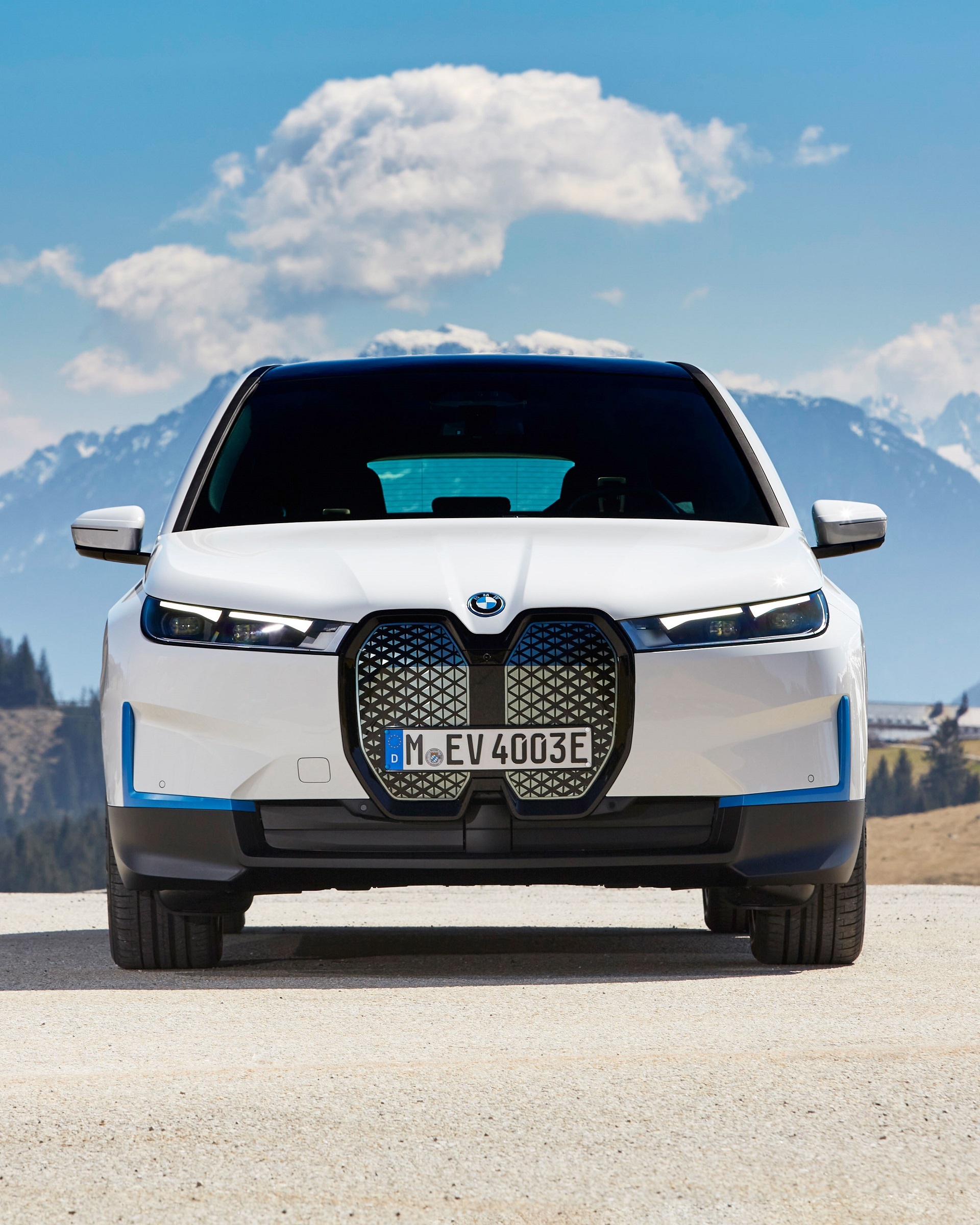 A white BMW iX electric SUV parked on a mountain road, with a backdrop of snow-capped mountains and blue sky