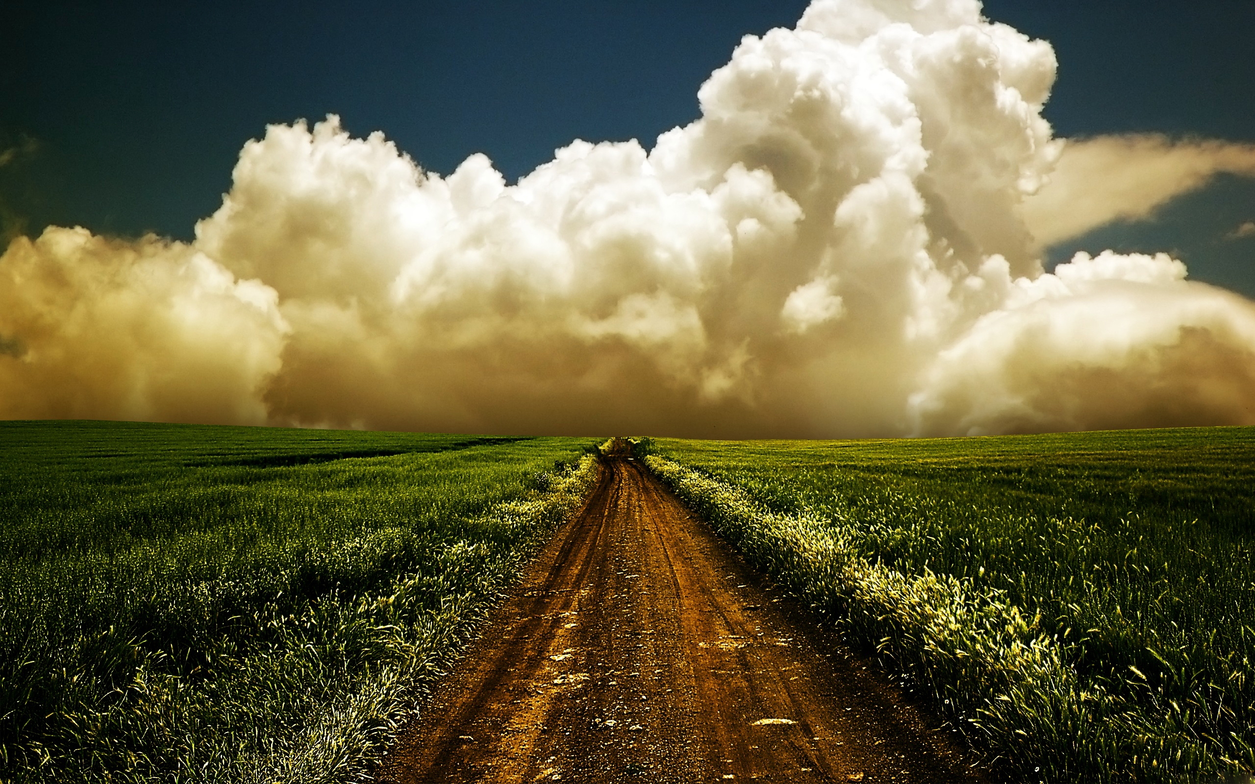 A dirt road cuts through a vibrant green field under a dramatic sky filled with large, illuminated cumulus clouds
