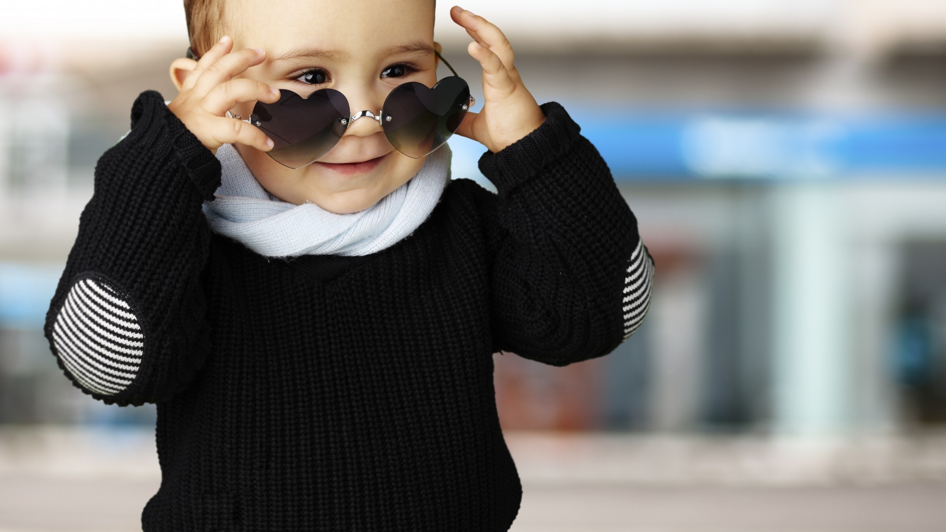 A stylish baby boy wearing heart-shaped sunglasses, a black sweater with striped elbow patches, and a light blue scarf